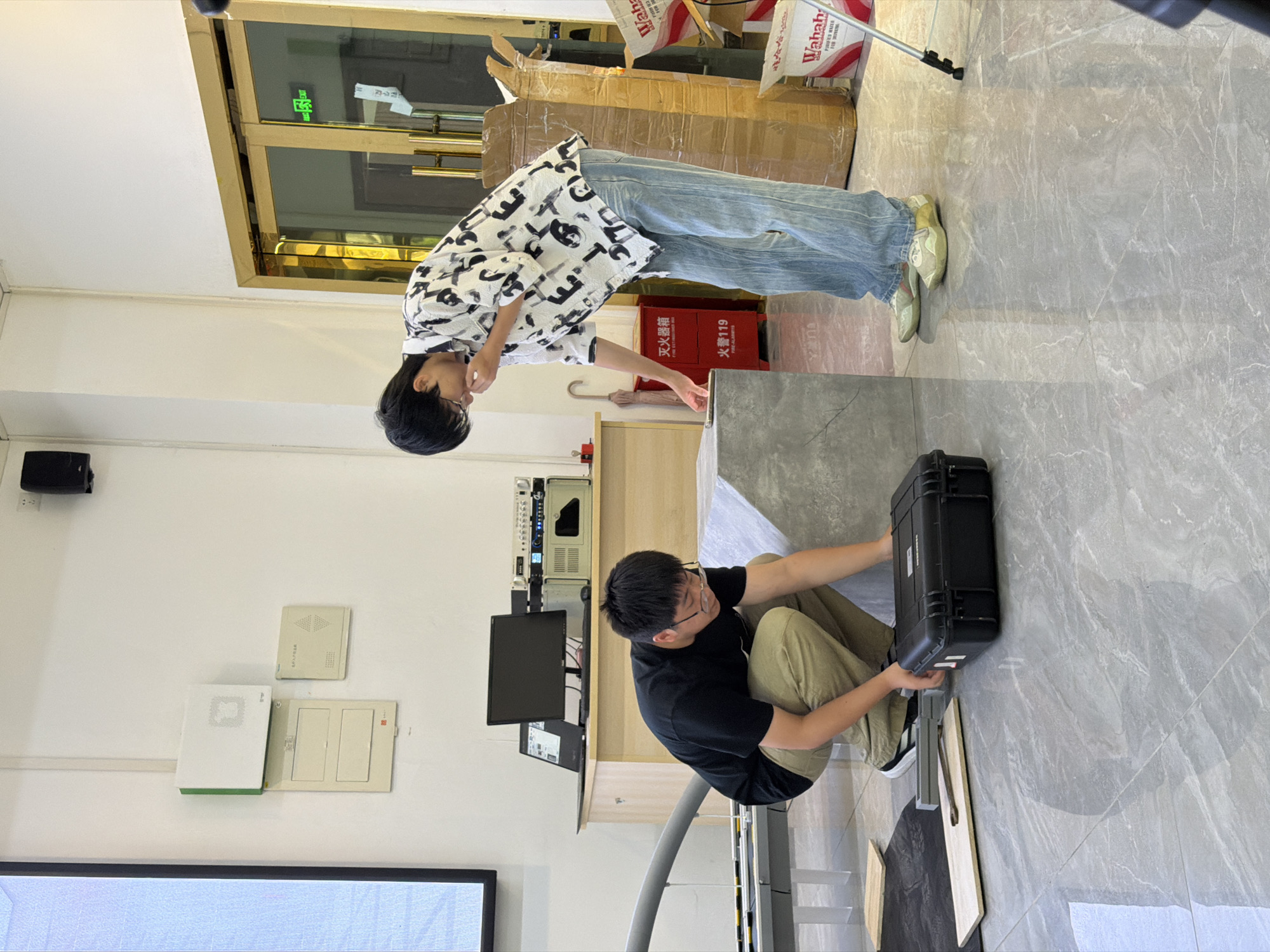 In a brightly lit indoor room with grey tiled flooring and white walls, two young men are engaged in an activity. The man on the left, wearing a black t-shirt and khaki pants, is crouched down examining a black hard-shell case on the floor. He is wearing glasses. To his right, a man in a patterned white and black shirt and light blue jeans is leaning forward, looking at a large concrete-like object. He is also wearing glasses. In the background, there is a desk with a laptop, monitor, and audio equipment. To the right, a large cardboard box is visible, along with a red fire extinguisher. The environment suggests a workshop or an event space. There are no specific visual cues to place the scene in Jiangjin, China.