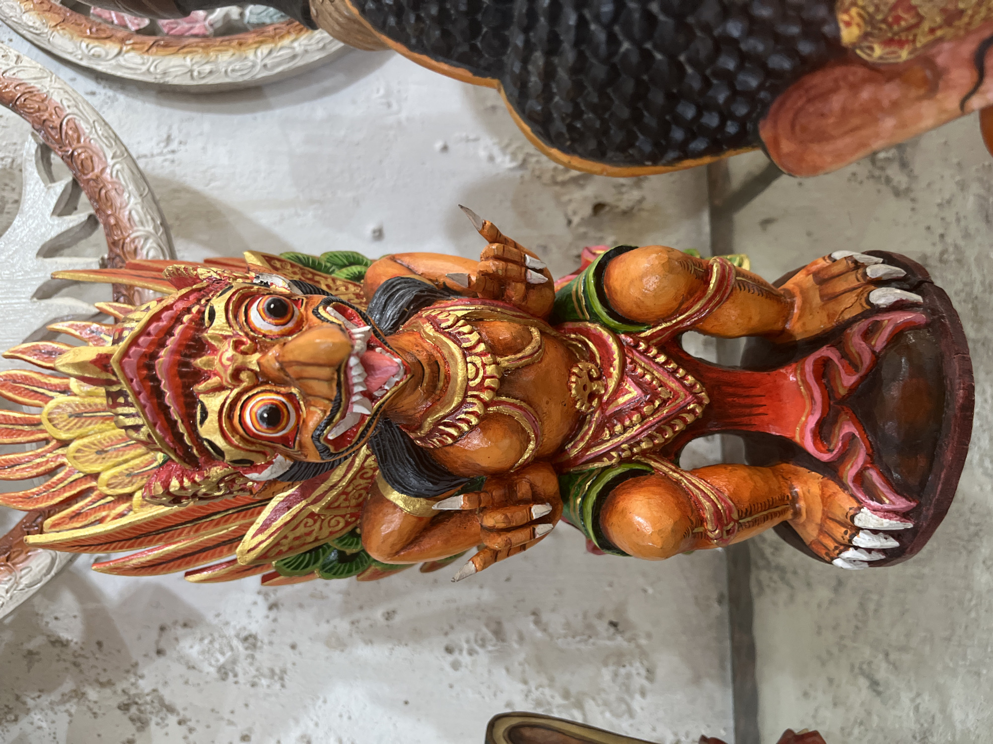 A close-up, medium-angle shot shows an ornately carved and painted wooden statue depicting a mythological creature, likely Garuda. The statue is primarily orange, red, and gold, with details in green, yellow, and black. The creature has a fierce expression with large, round eyes, sharp teeth, and clawed hands and feet. Decorative elements surround its body, including elaborate wings. The statue is surrounded by other carvings, indicating it is displayed in a shop or gallery. The location is most likely the Ubud District, Indonesia, which is known for its wood carving traditions and is a popular tourist destination. The statue is lying horizontally in the picture.