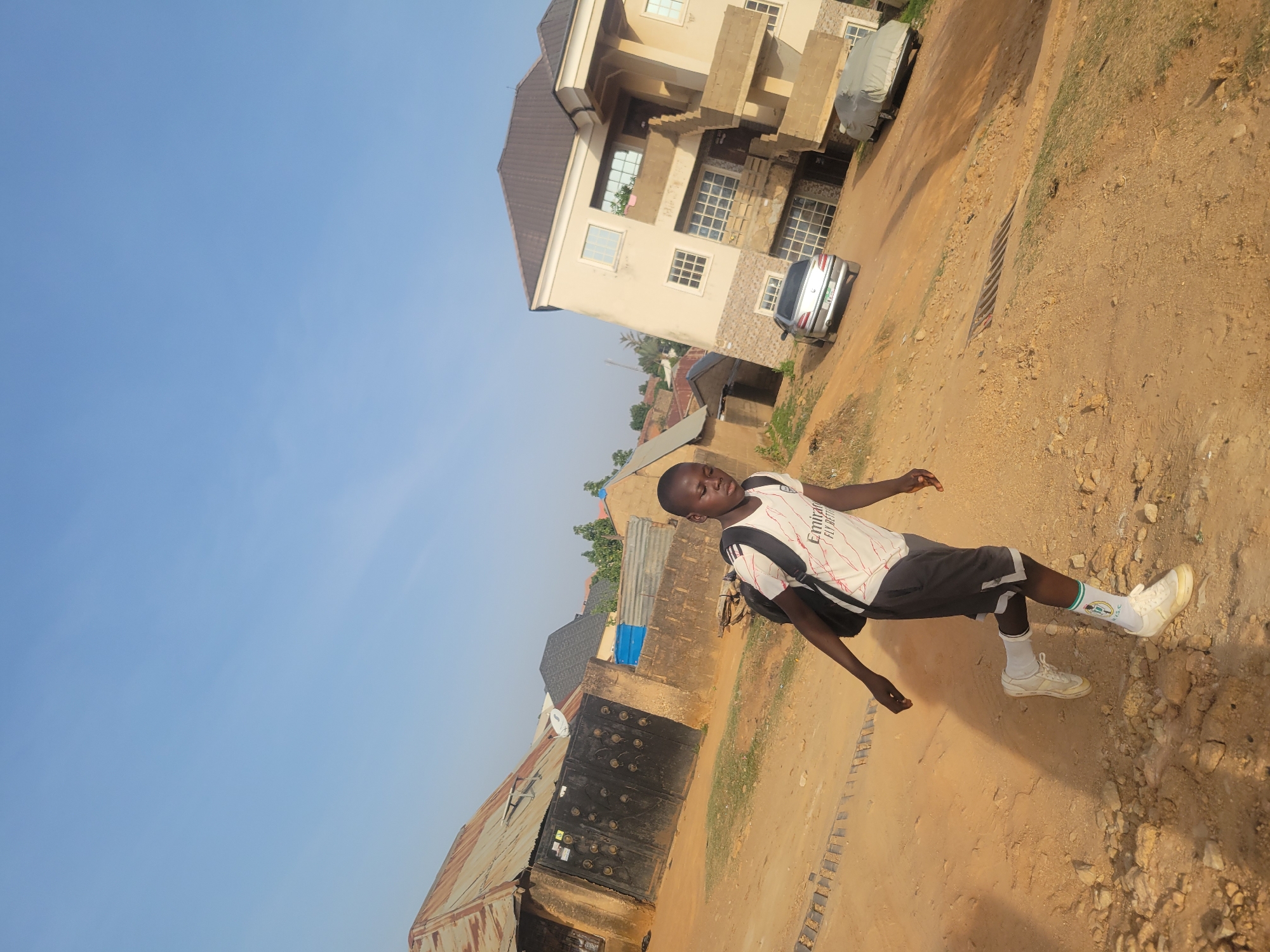 The image captures a young boy standing on a light brown dirt path in what appears to be a residential or semi-urban area of Jos, Nigeria. The boy, who is the central subject, has dark skin and short black hair. He is dressed in a white short-sleeved shirt with "Emirates FLY BETTER" printed in black on the front, dark shorts with white trim, white athletic socks (one featuring green and white stripes), and white athletic shoes. A black backpack is visible on his shoulders. He stands with his arms slightly out from his sides, looking towards his right.
The background features a mix of buildings and structures. To the left, there is an older building with a rusty corrugated metal roof and a dark, ornate metal gate. Further behind, other smaller structures are visible. To the right, a larger, multi-story building with a light-colored facade, numerous windows, and external staircases dominates the scene. Two cars are parked near this larger building; one is a silver sedan, and the other is covered with a grey tarp. A metal grate is also visible on the dirt path. The sky is a clear, bright blue, indicating good weather and daytime. The overall impression is a casual outdoor scene under clear daylight conditions.