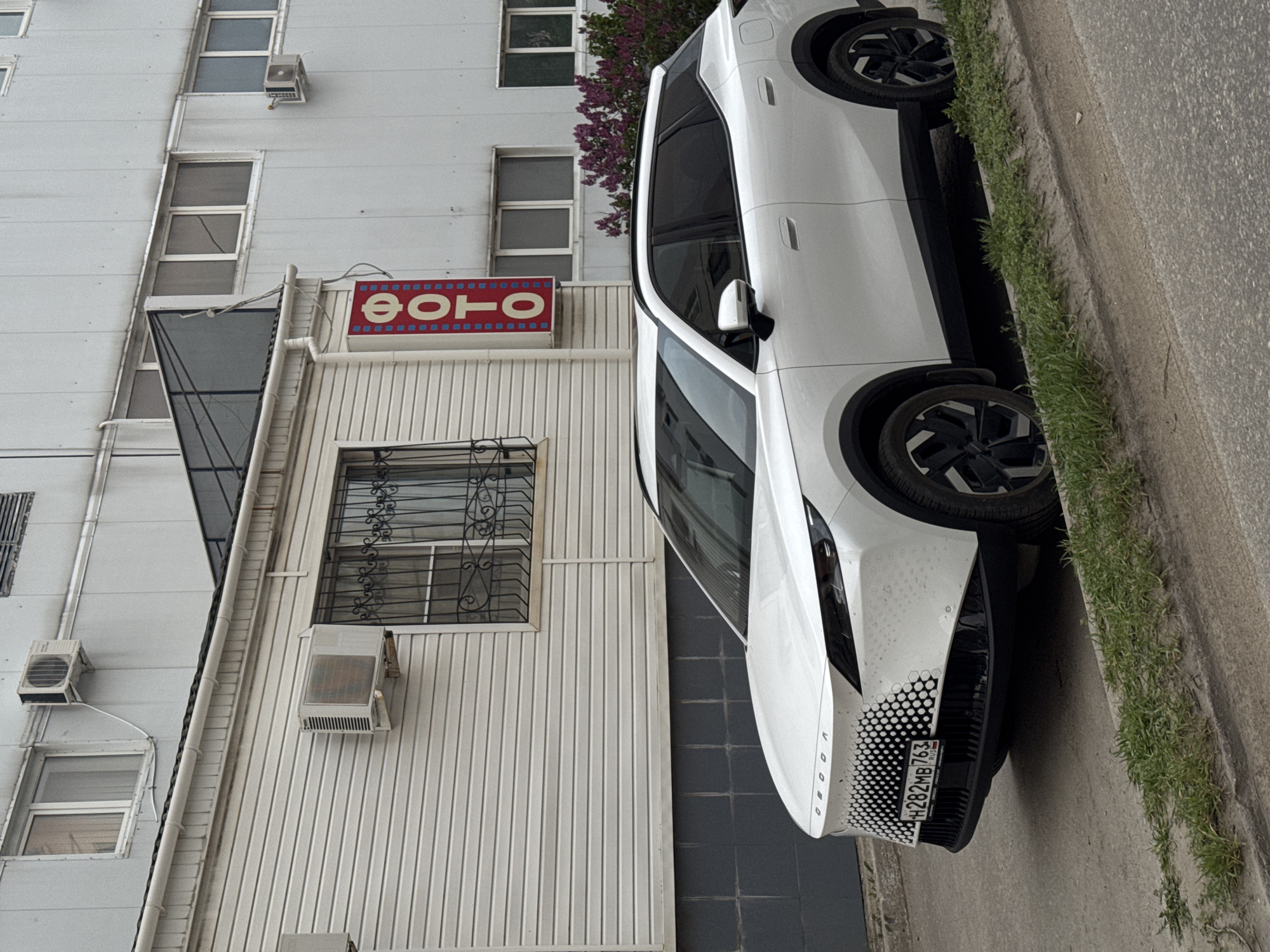 A white VOID electric car is parked on the side of a street in front of a building with a sign in Cyrillic script that reads "ФОТО" (photo). The license plate of the car reads "H282MB 763". The building has several windows, some with air conditioning units, and is painted in a light color. The sidewalk borders a curb with grass growing along its edge. The city is Unknown, Unknown.
