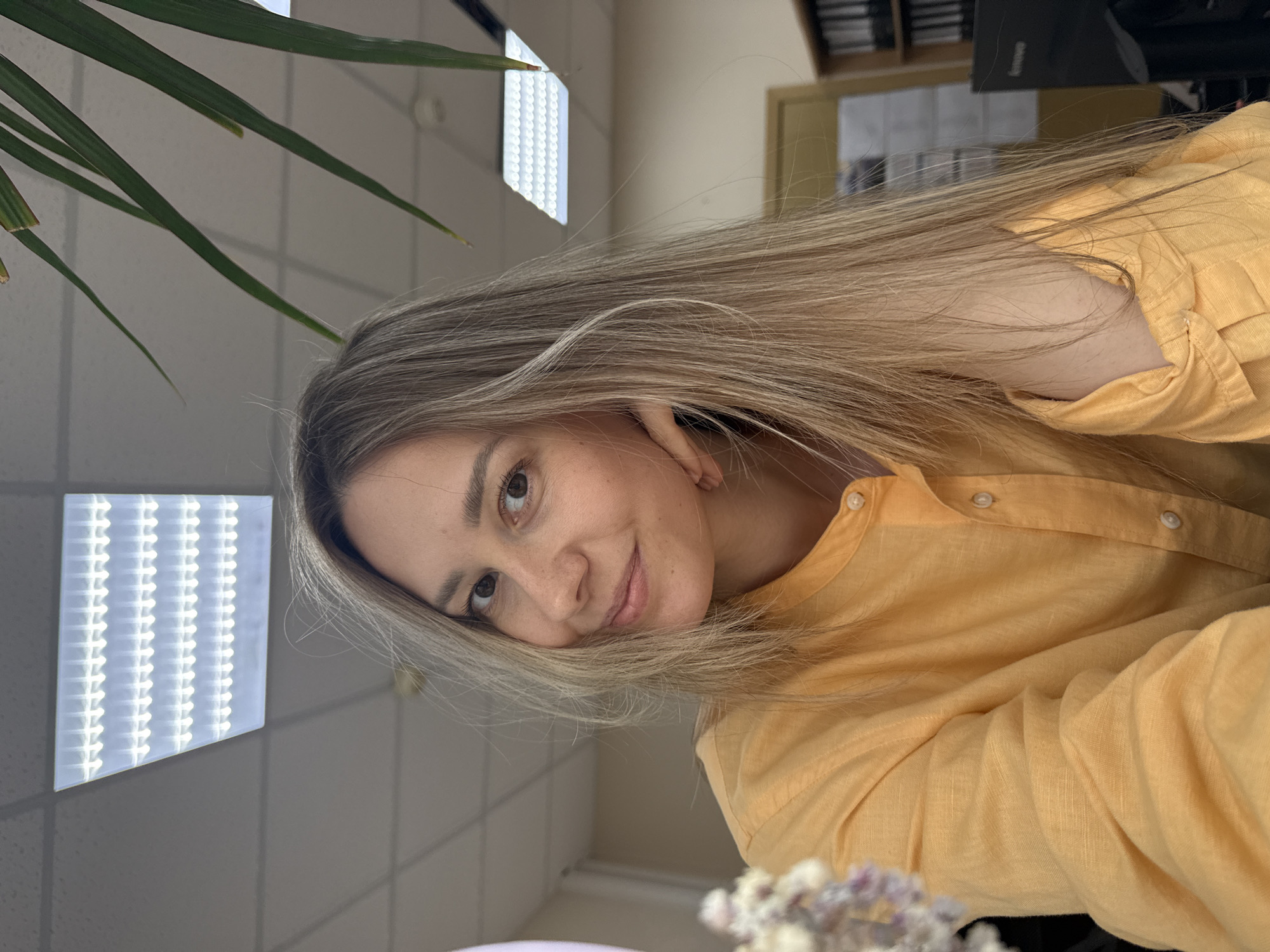 A selfie shows a fair-skinned woman with shoulder-length blonde hair wearing a yellow button-down shirt. She is smiling slightly and looking at the camera. The background includes a green plant in the upper left corner and a tiled ceiling with fluorescent lights. In the blurred background, there is a shelf filled with binders and a doorway. There are no indications of the specific location in the city of Chaikovsky, Russia.