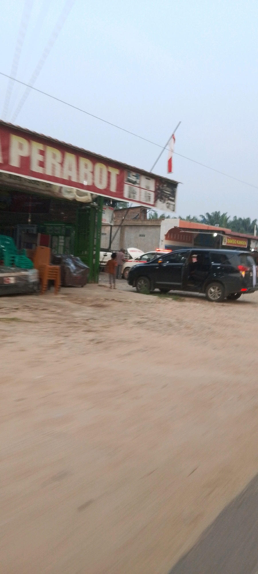 The image shows a storefront with a red and white sign that says "PERABOT" in large white letters. The store appears to sell furniture, with stacks of chairs visible inside and outside. A black SUV with its doors open is parked in front of the store. A white car with its hood up is parked next to the SUV, and a person in a red shirt is standing next to it. Another person in an orange shirt is standing in front of the store. In the background, there is a building with a sign that says "BAKSO KANGEN." The scene appears to be on a dirt road, with palm trees visible in the distance. The sky is overcast and the lighting suggests it is either early morning or late afternoon. The location is identified as Lenggadai Hulu, Indonesia.