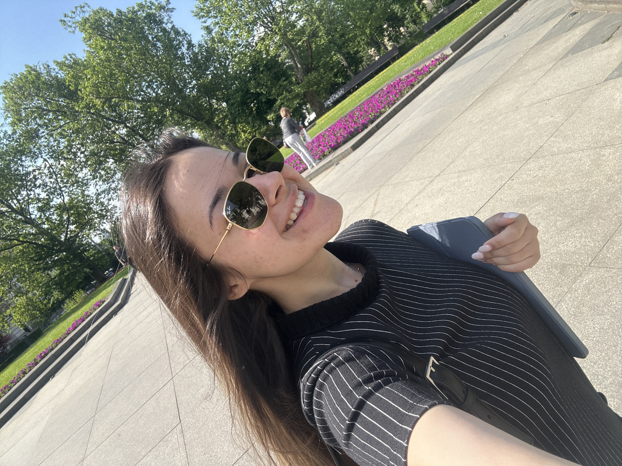 A young woman wearing sunglasses and a black and white striped shirt is taking a selfie outdoors. The background shows a public space, possibly a park, with trees and flowerbeds. A person is walking in the background. The location is most likely Sofia, Bulgaria, based on the architectural style and landscaping. The woman appears to be sitting down, as indicated by the visible chair and posture. The ground is paved, suggesting a plaza or walkway within the park.