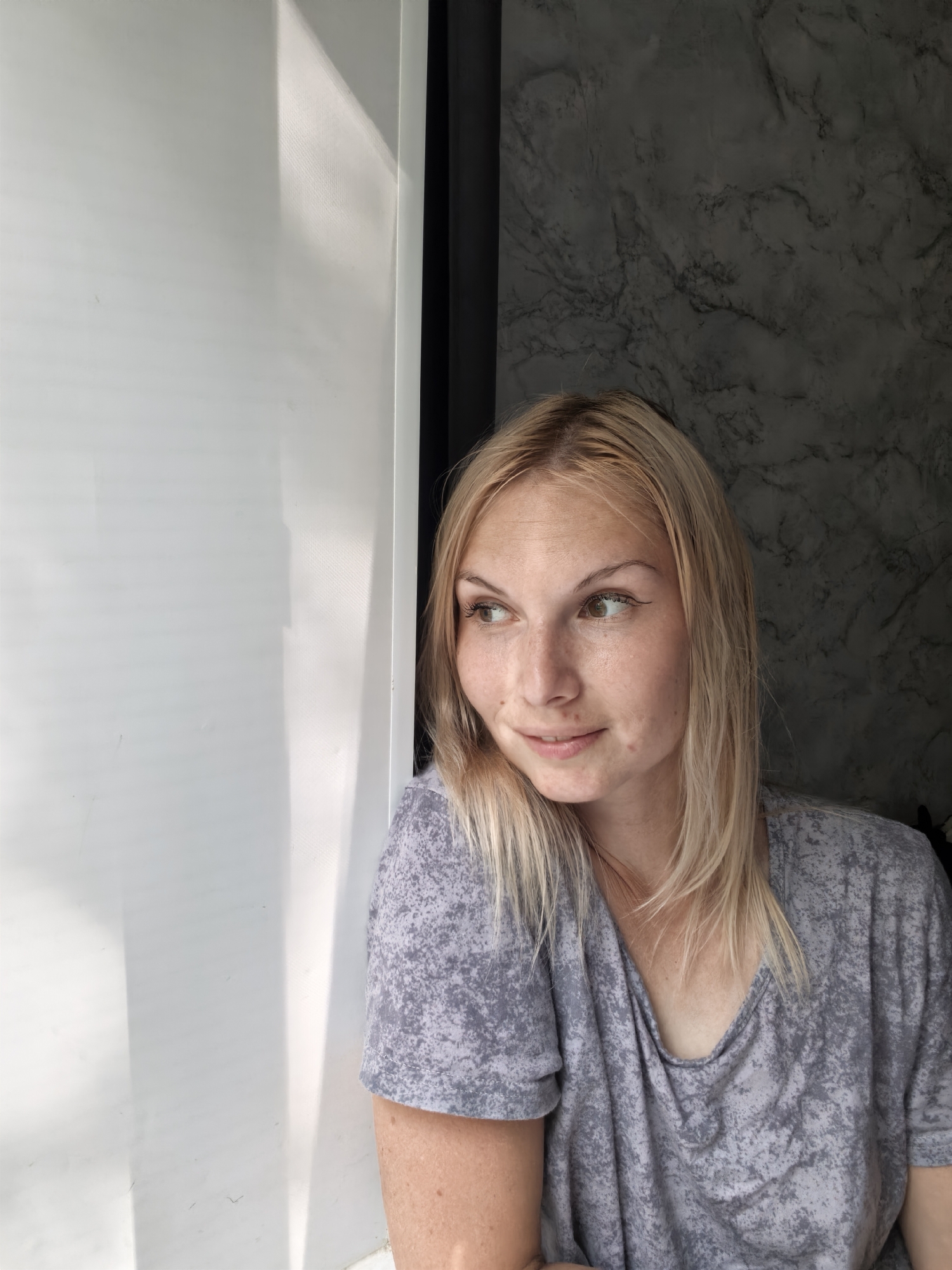 A young Caucasian woman with blonde hair and freckles is captured in a medium shot, looking to her left. She is wearing a grey t-shirt with a mottled pattern. The background features a stark white wall on the left, illuminated by natural light casting shadows, and a dark, textured wall with a marble-like pattern on the right. The scene appears to be indoors, possibly near a window or doorway, creating a contrast between light and shadow. The woman's expression is relaxed and contemplative. There is no indication of the location being in the city of Sterlitamak, Russia.