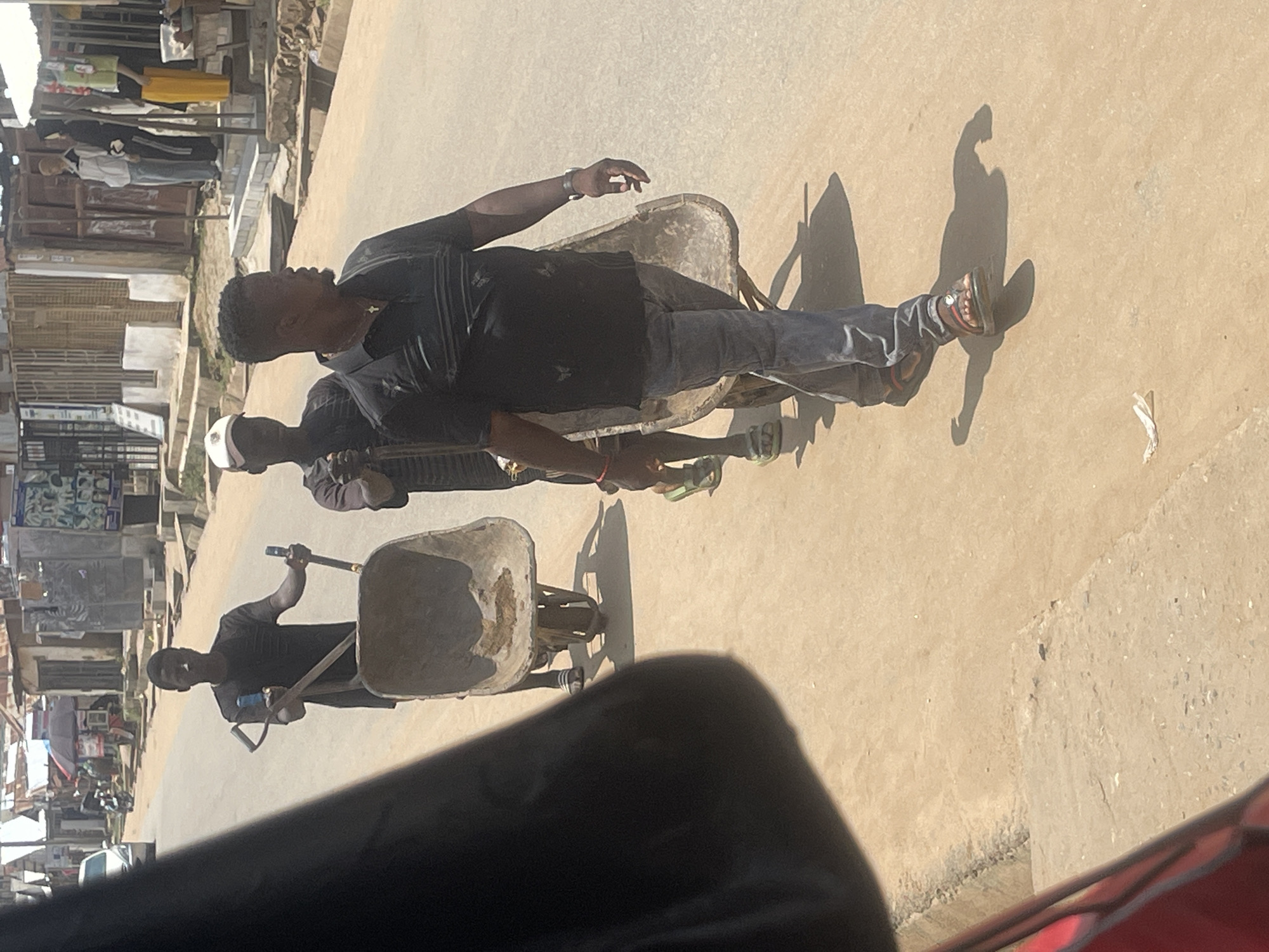 Three men are walking down a dusty, paved street in Abuja, Nigeria, each pushing a wheelbarrow. The man in the foreground, wearing a black polo shirt and blue jeans, walks to the right, looking straight ahead. He is pushing a wheelbarrow. To his left and slightly behind him, a second man in a dark shirt and white cap pushes another wheelbarrow. Behind him, a third man in a black t-shirt pushes a third wheelbarrow, walking to the right and looking ahead. The street is lined with buildings, some appearing to be shops, with various signs and displays visible. In the background, a white car is parked on the left side of the road. The sky is bright, suggesting a sunny day.