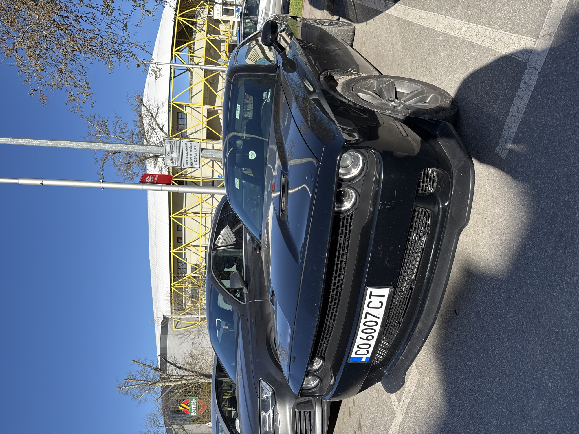 A black Dodge Challenger is parked in a parking spot in front of the "Botev" football stadium in Plovdiv, Bulgaria. The car has a Bulgarian license plate, "CO 6007 CT". There is a parking sign nearby indicating parking restrictions from 23:00 to 6:00. The sky is clear and blue.