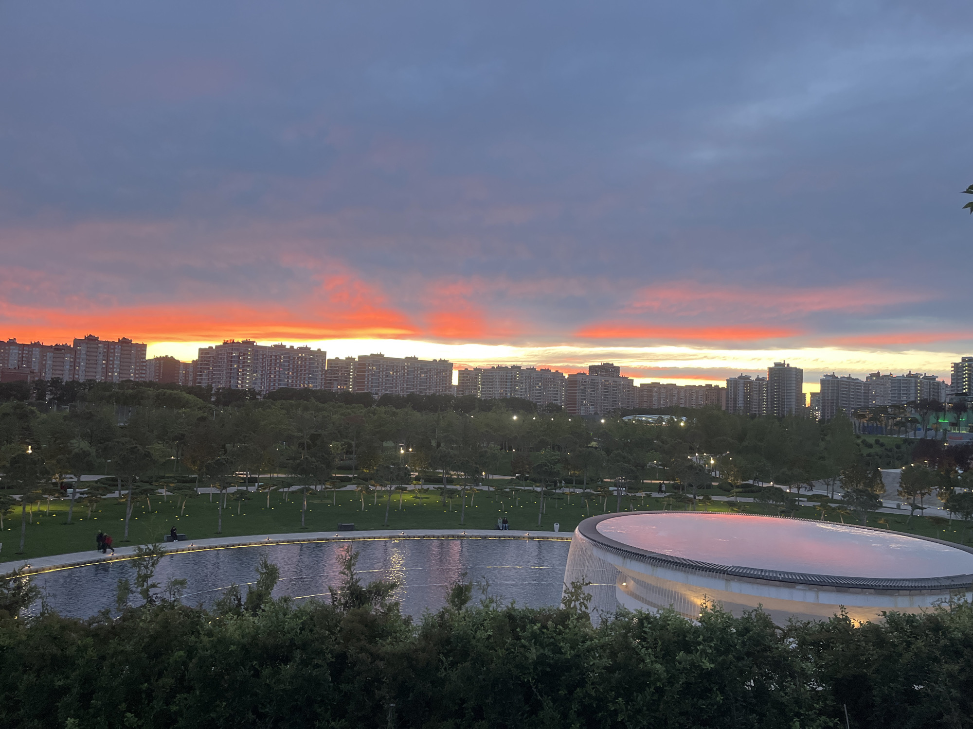 This image captures an expansive urban park scene in Krasnodar, Russia, at sunset. The sky is a prominent feature, transitioning from a dark grey-blue at the top to vibrant horizontal bands of bright orange and reddish-pink clouds along the horizon, creating a dramatic and colorful twilight display.
In the background, a dense array of modern, multi-story buildings forms a cityscape against the fiery sky. Many windows in these structures are illuminated, confirming the approach of evening.
The midground and foreground showcase a well-designed park featuring numerous trees, neatly maintained green lawns, and winding pathways subtly lit by ground-level lights. A large, curving water feature, likely a pond or decorative basin, lies in the foreground, its surface faintly reflecting the colors of the sky. Adjacent to this water body, on the right, is a distinctive, large, circular or oval-shaped modern structure with a light, reflective surface that also mirrors the sunset's glow, suggesting a pavilion or unique architectural element within the park.
A few people are visible enjoying the park's evening ambiance. Two individuals, one in a red coat, are seen walking along the pathway beside the water feature on the left. Another person is seated on a bench further along the path. Their presence suggests leisure activities such as strolling or relaxing. The overall scene evokes a sense of tranquil urban beauty, with the striking sunset providing a picturesque backdrop to the modern park and cityscape. There is no visible text in the image.