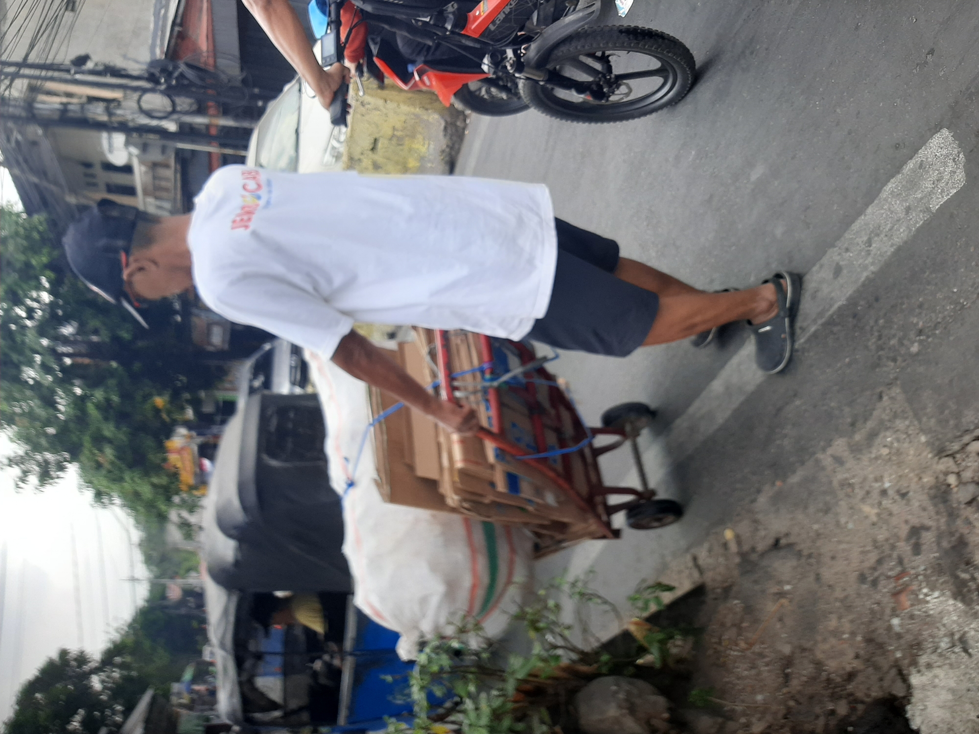 In Central Jakarta, Indonesia, on a cloudy day, a person is pushing a two-wheeled trolley laden with a large white bag and stacks of cardboard boxes down a paved street. The person, wearing a black baseball cap, white t-shirt with red and blue lettering, and dark shorts, is walking beside a red motorcycle. The scene is on a street with some vegetation on the left and buildings visible in the background, suggesting an urban or suburban environment. A three-wheeled vehicle, possibly a rickshaw, is partially visible in the background. The road has white markings.
