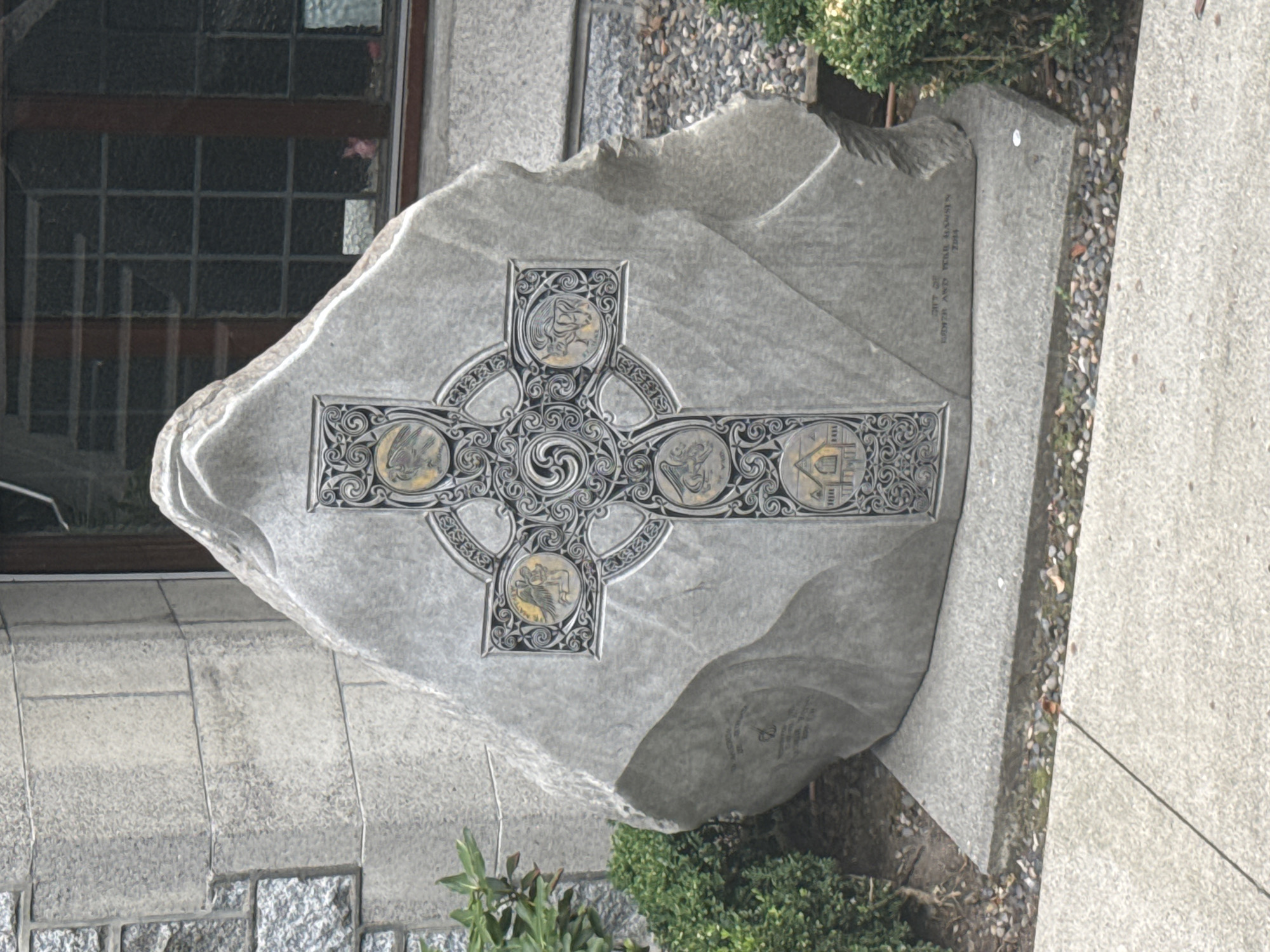The image depicts a large, rough-hewn stone monument with a Celtic cross design carved and inlaid into its face. The cross features intricate knotwork and circular medallions containing what appear to be figures or symbols, possibly representing religious or allegorical imagery. The stone itself is grey and textured, with visible striations and some natural imperfections. The monument rests on a flat, concrete base.
To the left of the monument is a patch of green foliage, likely a shrub or small tree. Behind the monument, a building wall made of large, rectangular stone blocks is visible, with a dark-paned window above. The overall setting appears to be an outdoor area adjacent to a building, possibly a church or a historical site.
The inscription on the lower right side of the monument is partially visible and reads "GIFT OF / BARBARA AND PETER HANSEN / 2014".
This monument is located at St. Michael's Church in Vancouver, Canada.