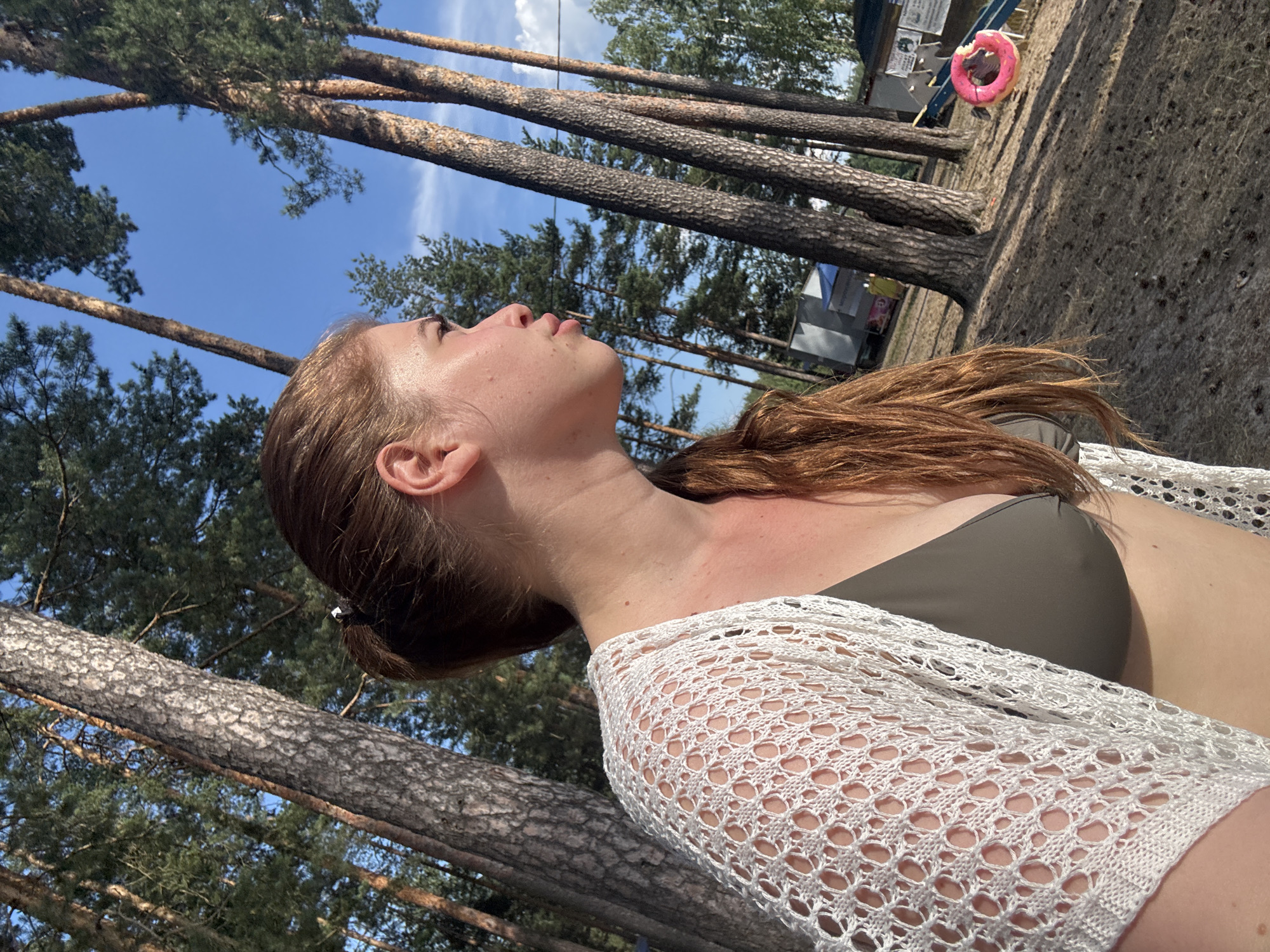 A young woman with auburn hair tied in a ponytail is looking to the right, away from the camera, with her profile visible. She is wearing a green bikini top and a white, crocheted, open-weave cardigan. The setting appears to be a forest clearing with tall pine trees. In the background, there are some structures, possibly temporary stalls or buildings, and a small pink ring buoy on the ground near a blue slide. The ground is covered in dry grass and pine needles. The sky is blue with a few wispy clouds. This scene is located in Kurovskoye, Russia.