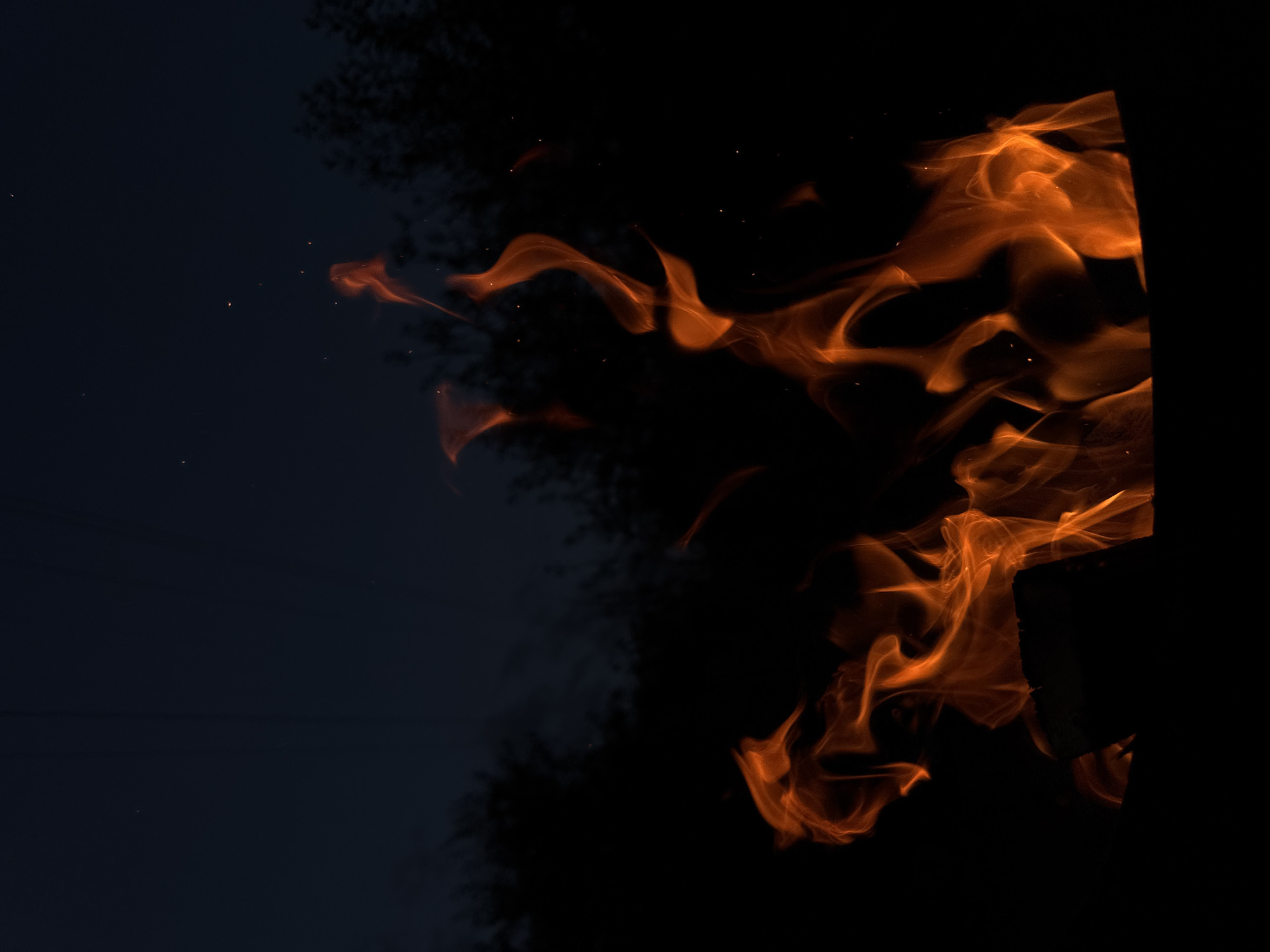 The image shows a close-up view of a fire burning against a dark background. The flames are a vibrant orange and yellow, with swirling patterns suggesting movement and heat. Several small, bright sparks are visible, indicating active combustion. The background is predominantly black, with some darker, shadowy shapes that could be trees or other structures silhouetted against the light of the fire. On the left side of the image, the background is a deep navy blue, transitioning to black towards the right. The contrast between the bright flames and the dark background creates a dramatic and intense visual. The overall impression is one of a contained fire, possibly a bonfire or a fire pit, set in a dimly lit outdoor environment. There is no visible text in the image. The image evokes a sense of warmth and energy, with a slightly mysterious or wild quality due to the darkness.
