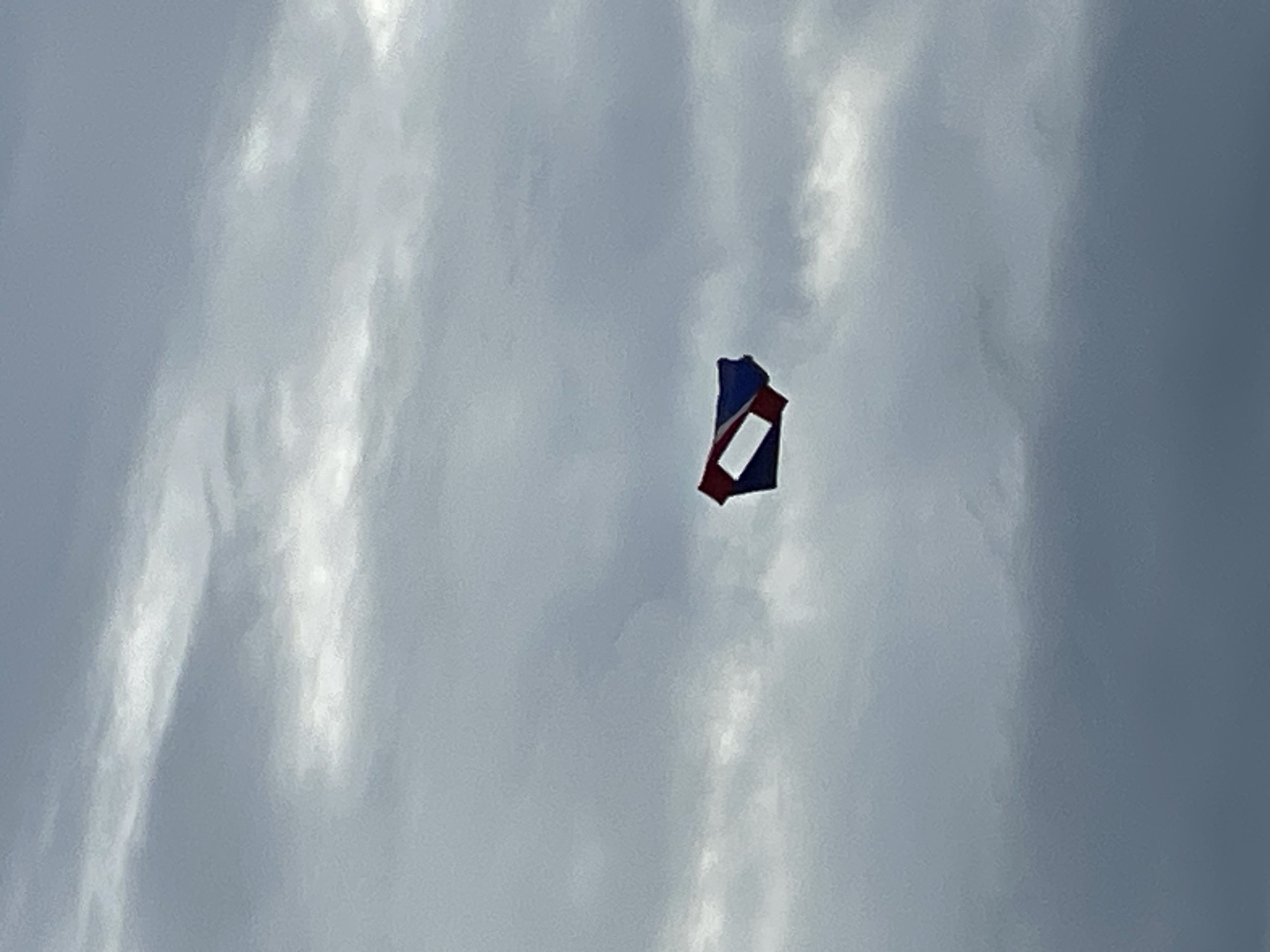 A single kite, appearing as a rhomboid shape with red and blue sections and a white rectangular opening, is visible against a backdrop of overcast, grey clouds. The kite is positioned in the upper right quadrant of the image and is the sole object of focus. The environment is a sky without any discernible city landmarks, urban features, or signs of human presence, making it impossible to determine its location within Pattaya City, Thailand.