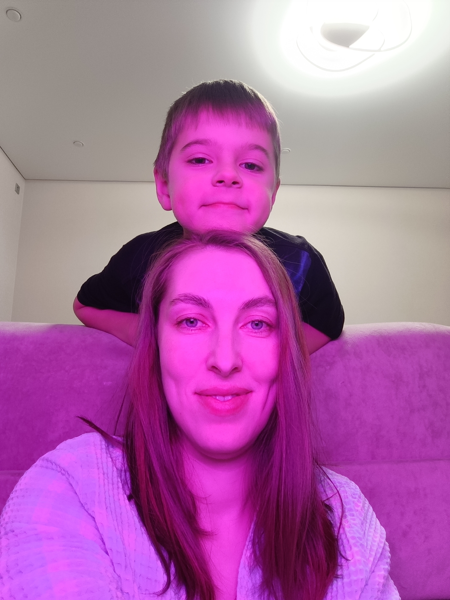 An indoor portrait captures a woman and a young boy in a modern living space in Chaikovsky, Russia. The woman, positioned in the foreground, faces the camera with a subtle smile. She has long, straight hair, which appears magenta due to the dominant pink and purple ambient lighting, and is wearing a light-colored, textured garment. Behind her, a young boy leans over the backrest of a couch, also looking towards the camera with a slight smile. He wears a dark t-shirt, and his face and hair are similarly illuminated with a pink-purple glow.
The setting features light-colored walls and a plush, textured couch, which also takes on the pink-purple hue of the lighting. Overhead, a modern, bright circular ceiling light fixture emits white light in the upper right, contrasting with the colored light. Additional recessed ceiling lights are visible in the upper left. The photograph appears to be a casual selfie, likely taken in the evening or at night given the reliance on artificial lighting. Both subjects display relaxed and pleasant emotions. No text is visible in the image.