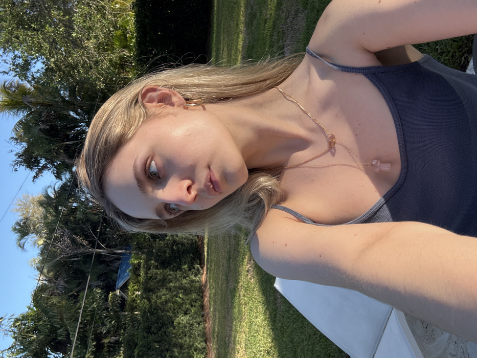 A young Caucasian female is depicted in a close-up outdoor self-portrait, seated or reclining on a white lounge chair in a residential setting in Coral Gables, United States of America. She possesses long, straight blonde hair and is looking upwards and to her left. Her visible attire includes a dark grey or blue ribbed tank top, gold-colored hoop earrings, a short gold-colored necklace with interlocking ring pendants, and a longer gold-colored necklace with a light pink mushroom-shaped pendant. The scene is illuminated by natural sunlight, casting a warm glow on her face. The background consists of a manicured green lawn, dense green hedges, and numerous tall palm trees and tropical foliage under a clear blue sky. A utility line is faintly visible across the upper left sky. A structure with a blue roof is partially obscured by trees in the distant background.