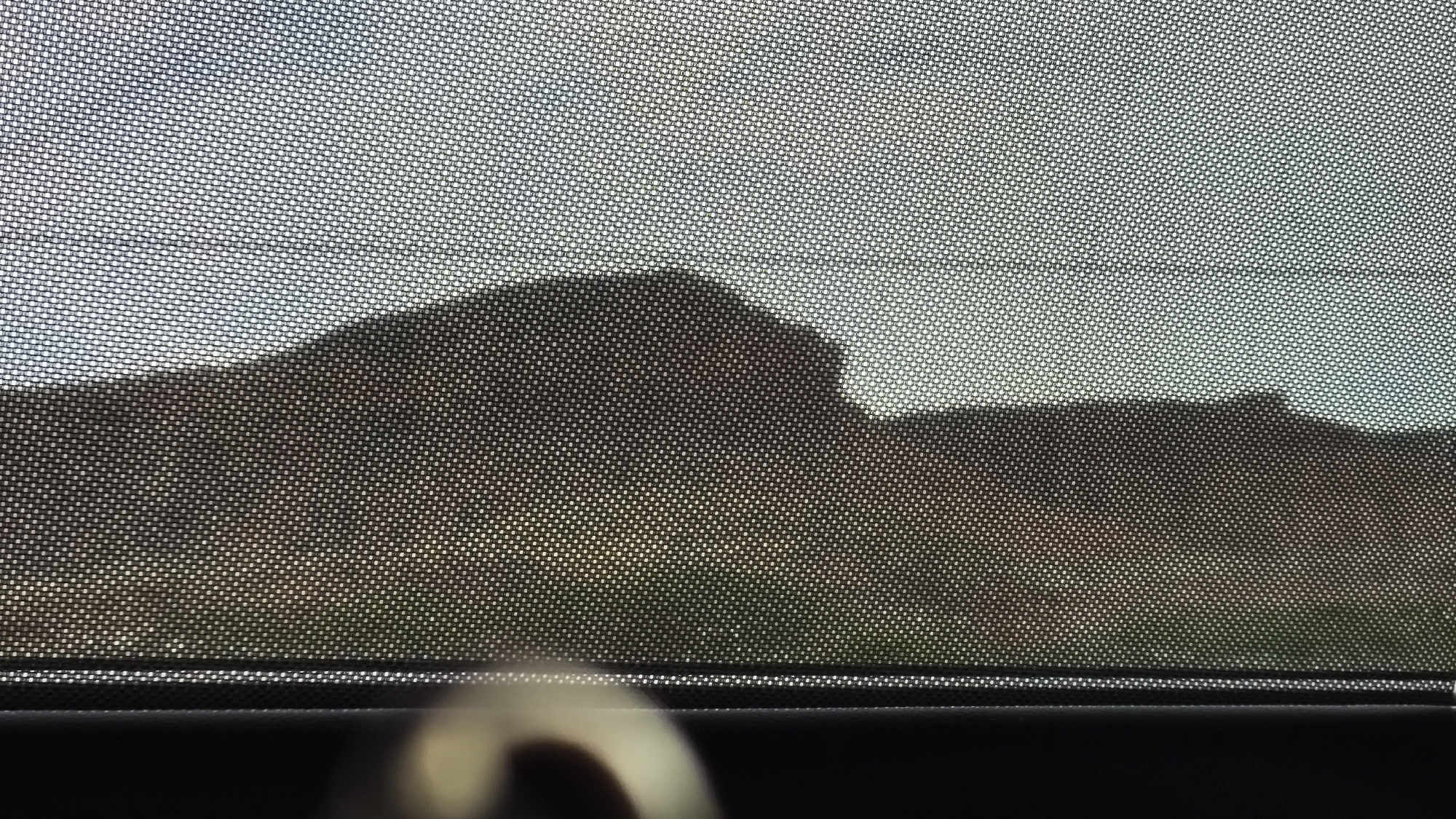 The image, taken from inside a vehicle, shows a sunshade covering the window. Through the mesh of the sunshade, a silhouette of hills or mountains is visible against a bright sky. The lower part of the image shows the interior of the car, with a dark surface and a blurred circular object, possibly a person's hand or a camera lens. The scene does not provide any specific visual cues to determine its location within the city of Rockville, United States of America.