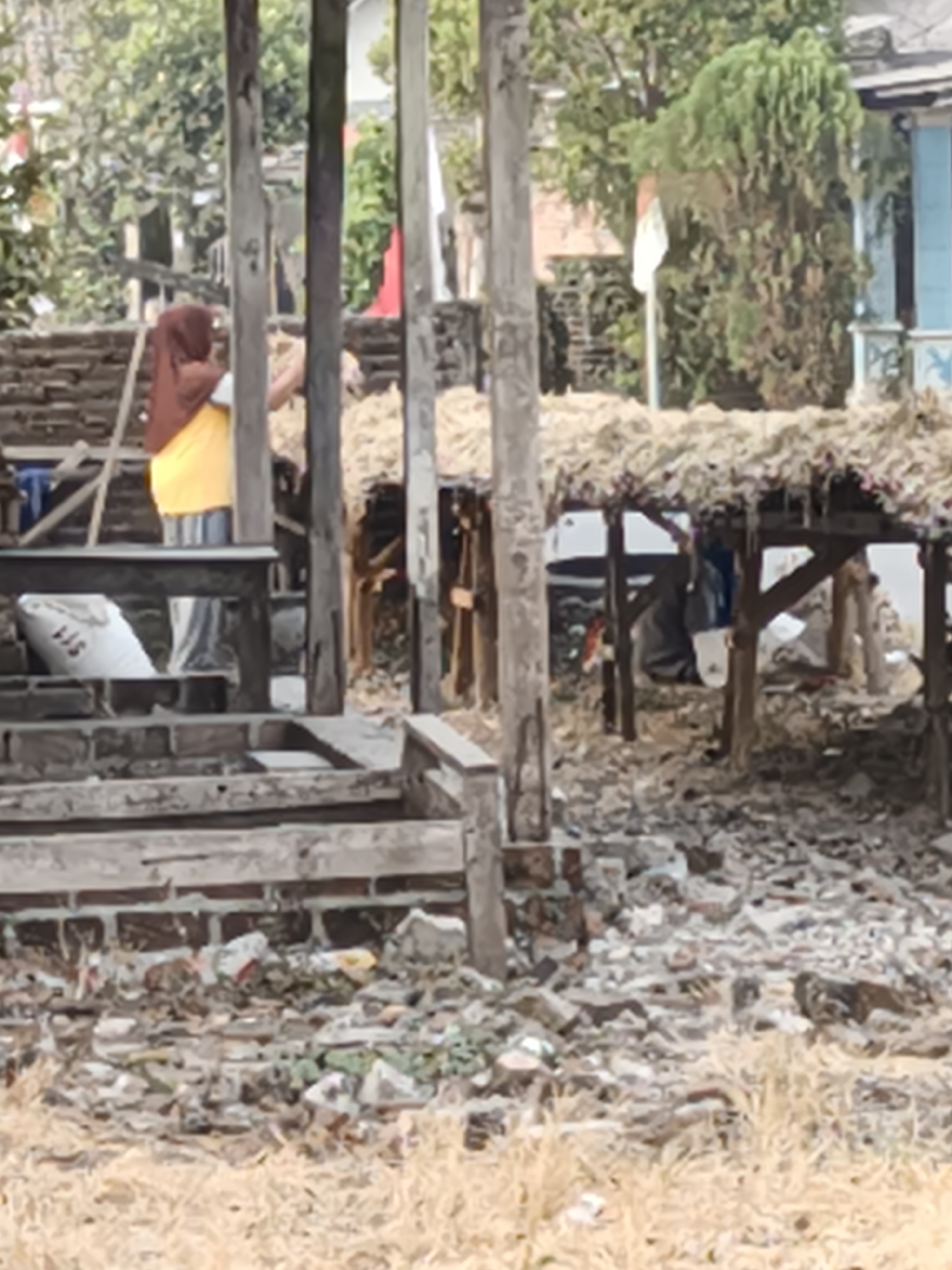 The image shows an outdoor scene in Pandean, Indonesia, characterized by a rustic and somewhat dilapidated environment. In the foreground, there is a pile of debris, including broken bricks and what appears to be dried vegetation or straw. A rough wooden structure, possibly a stall or shelter with a thatched roof, stands in the mid-ground, supported by wooden posts.
To the left of this structure, a person wearing a dark headscarf and a yellow garment is visible. They are standing near a wooden bench or table, on which a white bag with some markings is placed. Behind them, a brick wall is partially visible.
Further into the background, more of the thatched-roof structure is seen, with shadows suggesting it is open on at least one side. The general impression is one of a rural or semi-rural setting, potentially a market area or a humble dwelling, with an emphasis on natural and salvaged materials. The presence of what could be Indonesian flags in the background suggests a possible connection to national holidays or events. The overall lighting is diffused, indicating an overcast day or shade.