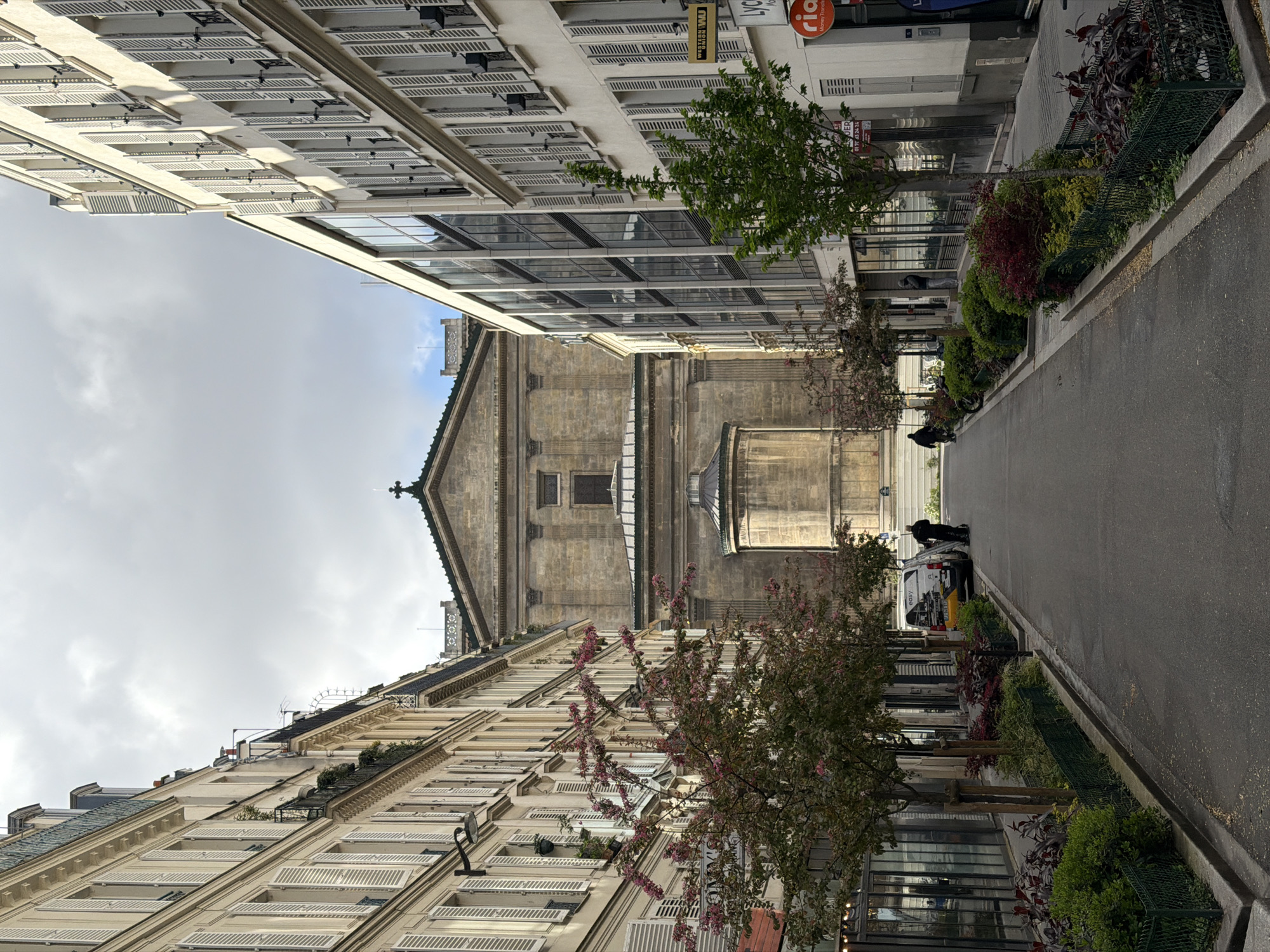 The image depicts a street in Paris, France, lined with buildings on both sides. The buildings feature classic Parisian architecture, with multiple stories, windows with white shutters, and ornate details. A church structure is visible at the end of the street. The street itself is paved and has greenery planted along the edges, including trees and shrubs. There are some people walking on the street, and a white van parked. There are signs for "easy" and "ria" visible on the buildings. The sky is overcast.