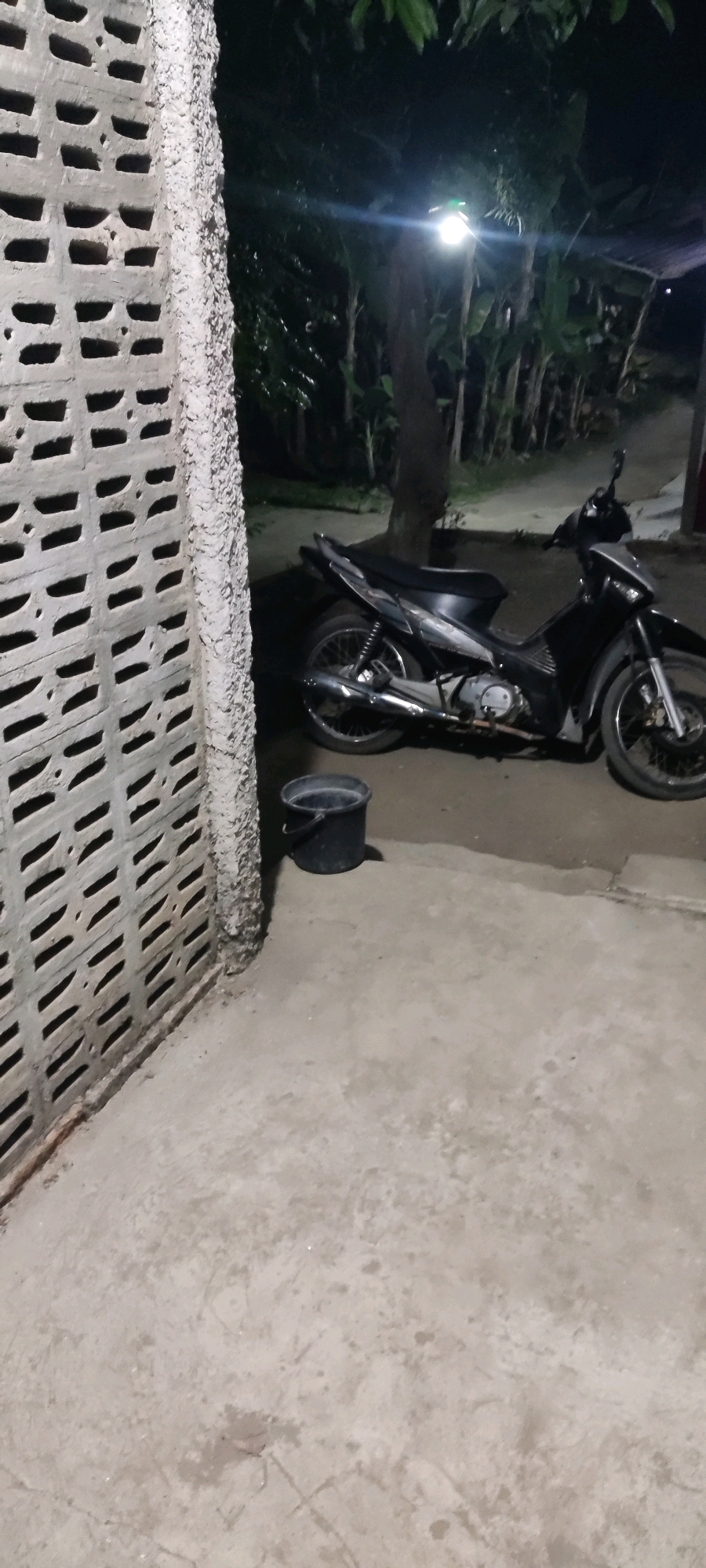 The image depicts a nighttime outdoor scene in Karangwangun, Indonesia. In the foreground, a concrete surface is visible, textured with faint lines and occasional dark spots. To the left, a portion of a white concrete wall with a regular pattern of rectangular openings runs vertically along the frame. Adjacent to this wall and slightly to the right, a black plastic bucket sits on the concrete. Behind the bucket, a black motorcycle is parked. The motorcycle is angled, with its front wheel facing towards the right and its rear wheel towards the left. It has a silver accent on the front panel and a black seat. The background is dark, with foliage and trees visible, suggesting a somewhat rural or vegetated area. A bright light source, possibly a lamp, illuminates the upper right portion of the scene, casting a glare. The ground appears to be a mix of concrete and dirt.