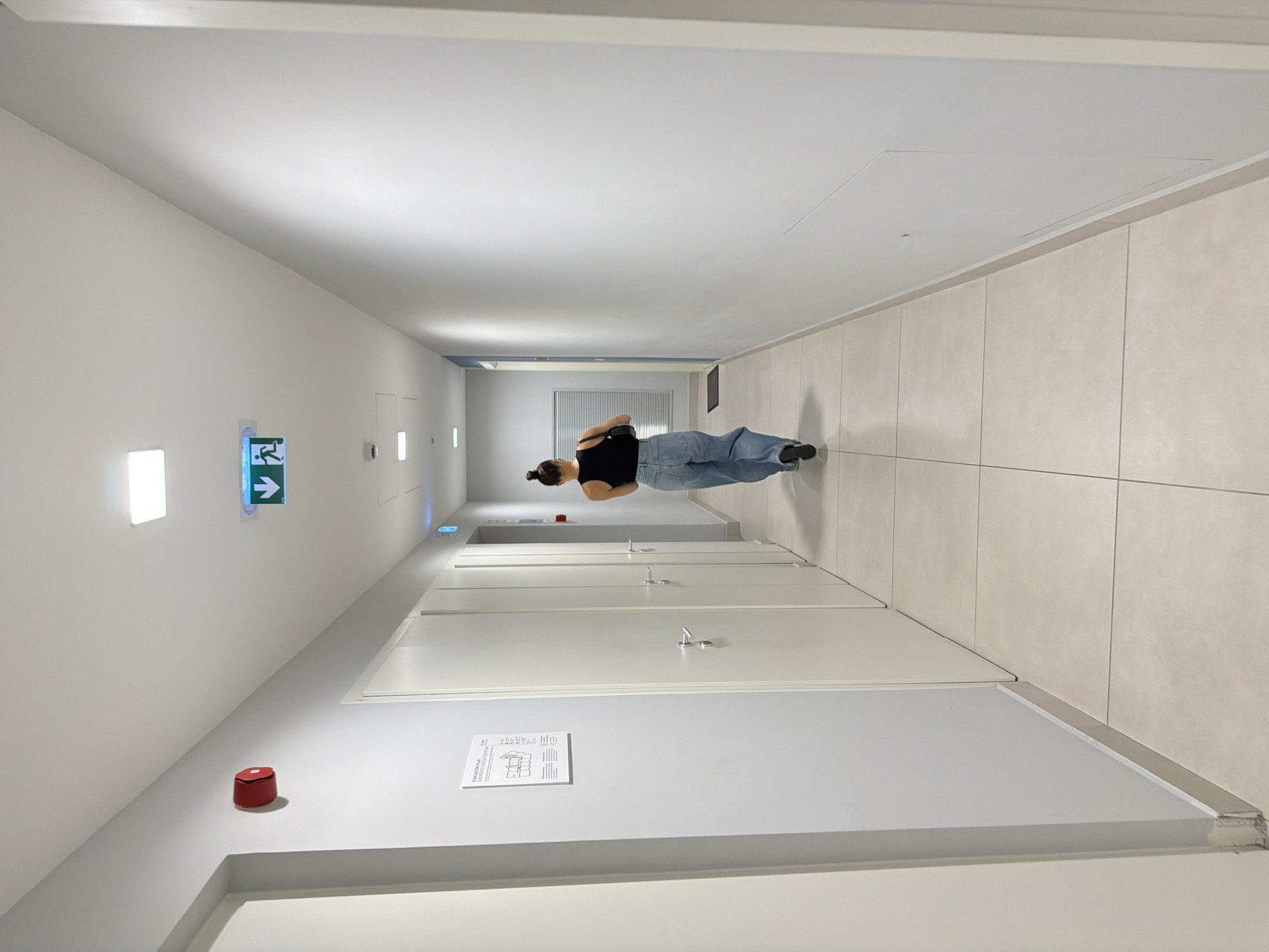 A woman walks down a corridor with tiled floors and white walls. The corridor appears to be indoors, possibly within an office building or apartment complex. Exit signs are visible on the left wall, along with a fire alarm pull station and emergency evacuation plan. The location is potentially Belgrade, Serbia.