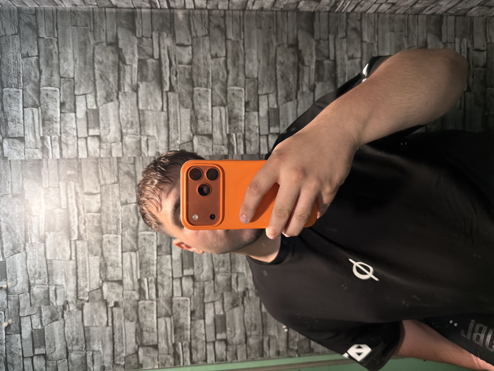 A male individual is captured in a mirror selfie taken indoors. He is seen from the chest up, with his head tilted slightly to his left. He has short brown hair and light facial stubble or a short beard. He wears a plain black t-shirt adorned with a white circular symbol featuring a diagonal line through its center, located on the left side of his chest.
The man holds an orange smartphone, likely an iPhone given its triple camera array, with his left hand. The phone is positioned to take the photo, partially obscuring the lower half of his face. The background consists of a grey wall with a textured pattern resembling stacked stone or brickwork, suggesting it might be wallpaper or a decorative panel. A bright light source emanates from the left side of the image, casting a glow on the wall. Below the man, the reflective surface of the mirror shows a blurred view of his lower torso and possibly reversed letters "BT" on a dark strap or garment.
The setting is an indoor environment, located in Sofia, Bulgaria. The time of day is not explicitly discernable, but the lighting suggests an interior space with a prominent light source. No strong emotions are visible on the man's partially obscured face.