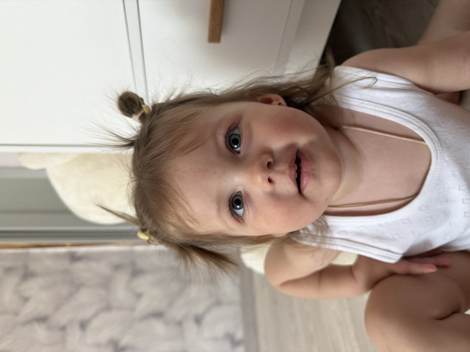 A child is looking directly at the camera. The child has fair skin, light brown hair, and blue eyes. The child's hair is styled in small pigtails on top of their head. The child is wearing a white tank top with a thin strap necklace. The background includes a white cabinet and a patterned floor. The image does not contain information to determine its location in Reshetnikovo, Russia.