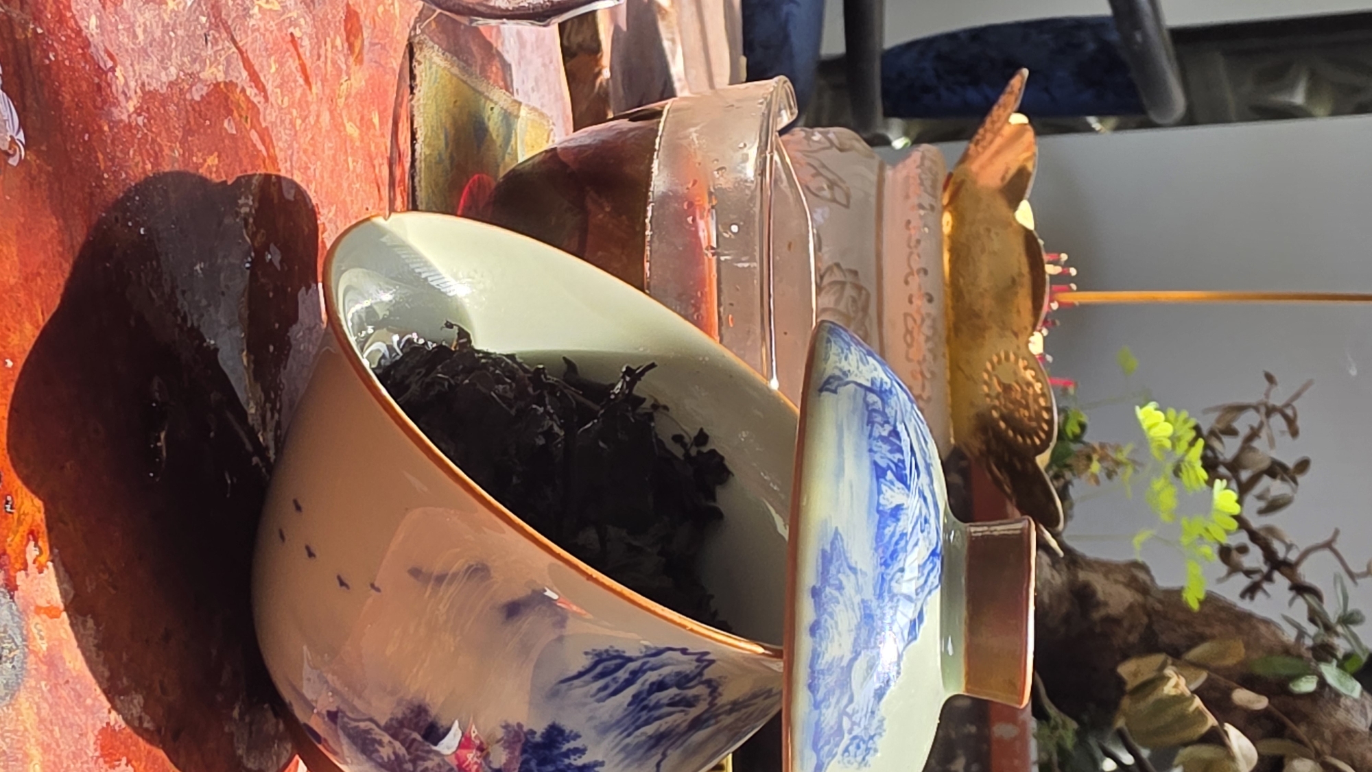 The image, taken in Bac Nha Trang Ward, Vietnam, captures a close-up of a ceramic teacup (gaiwan style) with its lid, presented at an angle. The teacup is exquisitely decorated with traditional blue and white landscape patterns, featuring mountains and a scattering of small bird-like designs. Inside the cup, dark, loose tea leaves are visibly steeping in water, indicating a tea preparation process.
Strong, directional sunlight illuminates the scene, creating prominent, dark shadows of the teacup and its lid on the textured, reddish-orange surface behind them. This background surface appears somewhat mottled or painted. Bright reflections of the light are visible on the water within the cup and on the glossy ceramic.
In the upper right background, a golden-brown, intricately patterned decorative object, possibly a stylized butterfly or bird, stands vertically. Behind this, subtle glimpses of green foliage from a plant or artificial arrangement emerge, alongside a darker, gnarled, branch-like element. A segment of blue upholstered furniture or a chair is partially visible in the far upper background. The scene conveys a tranquil moment of tea brewing, bathed in natural light. There is no visible text in the image.