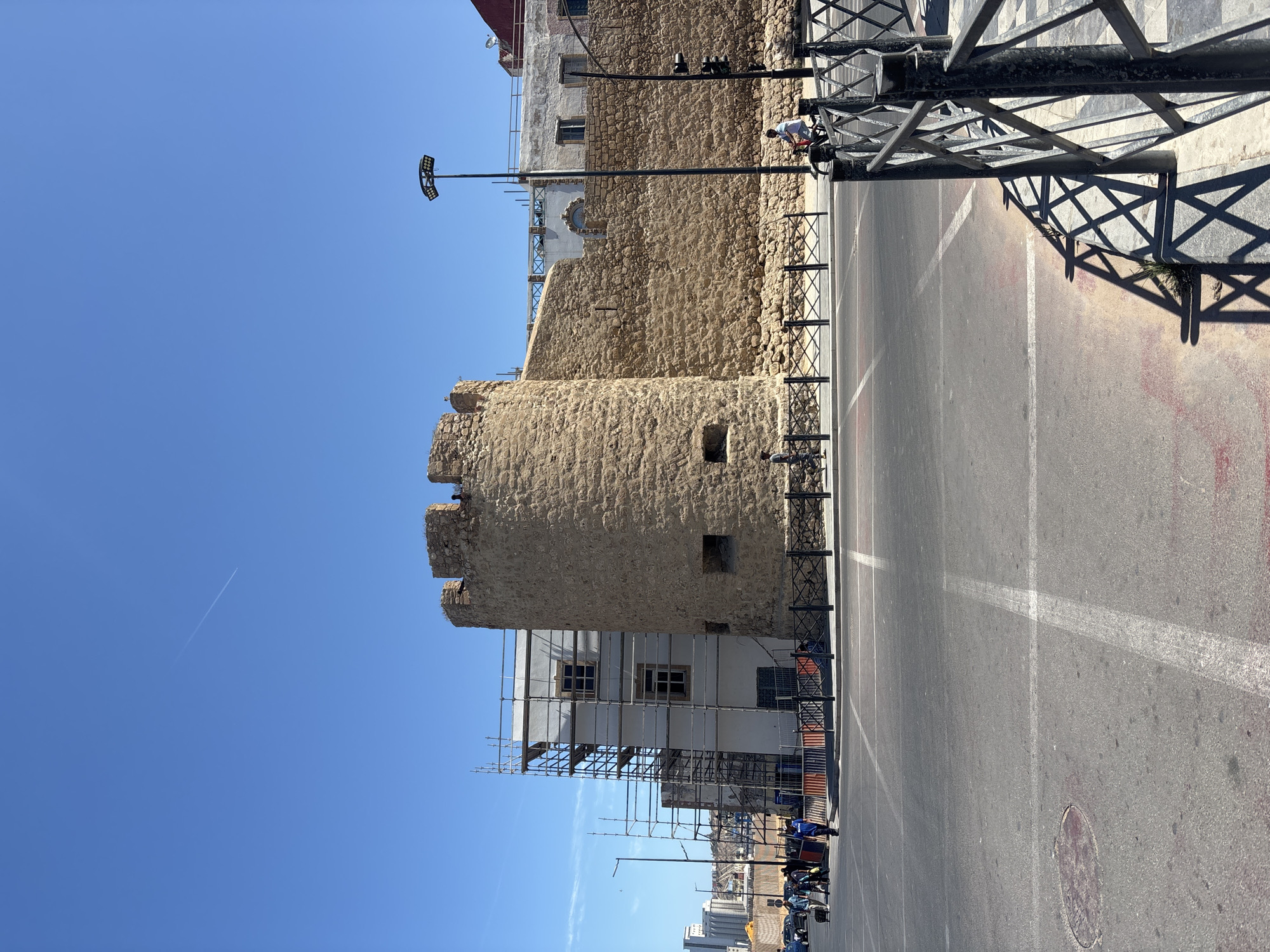 The image depicts a segment of an ancient stone wall and a partially visible modern building under construction with scaffolding, set against a clear blue sky. The stone structure, a prominent tower and connecting wall, appears weathered and old, with small rectangular openings and a crenelated top. Adjacent to it, a multi-story building with white walls and windows is covered in scaffolding, indicating ongoing renovation or construction.
In the foreground, a paved street with white lane markings and crosswalks is visible. Several people are seen on the sidewalk behind a black metal fence. Some individuals are walking, one is pulling a wheeled suitcase, and another is seated. A person on a bicycle is also present on the street, heading away from the camera. A tall lamppost with multiple lights stands near the stone wall. Traffic lights are visible in the distance. The environment appears to be an urban setting, likely a city square or a street adjacent to historical fortifications. The lighting suggests it is daytime with bright sunlight, casting shadows from the fence and the lamppost.