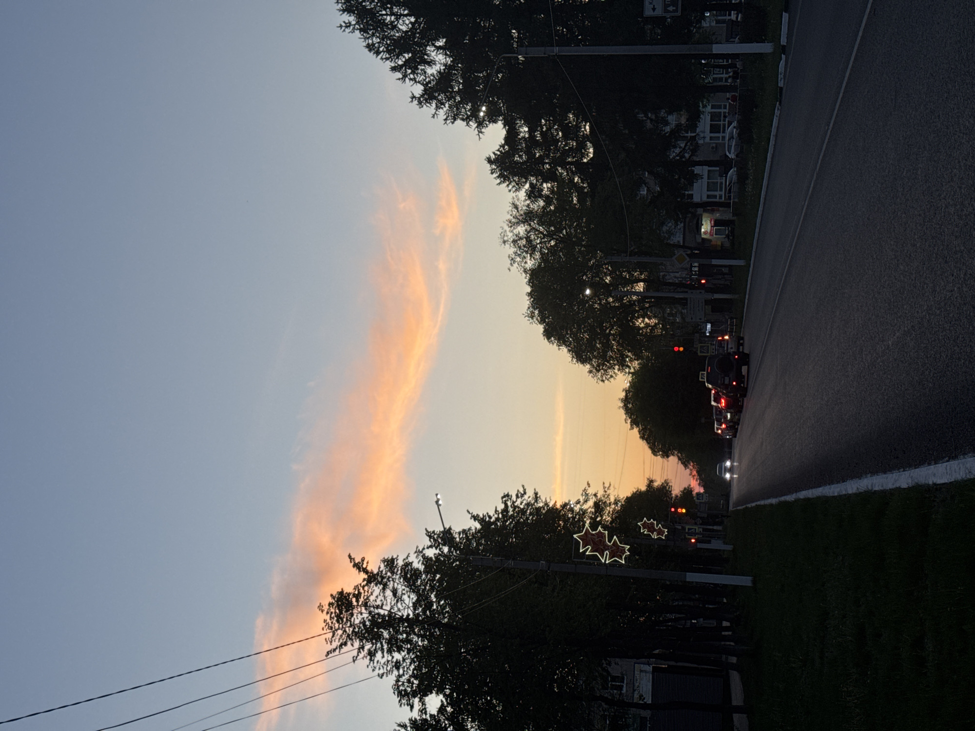 The image depicts a street scene in Chaikovsky, Russia at twilight. The sky is a gradient of blue to orange, with streaks of pink clouds. A road runs along the right side of the frame, with parked cars and traffic visible further down the street. Trees line both sides of the road, obscuring buildings and shops. Street lights are on, including some adorned with decorative star-shaped lights. Power lines run along the left side of the frame. The overall impression is of a quiet evening on a typical city street.