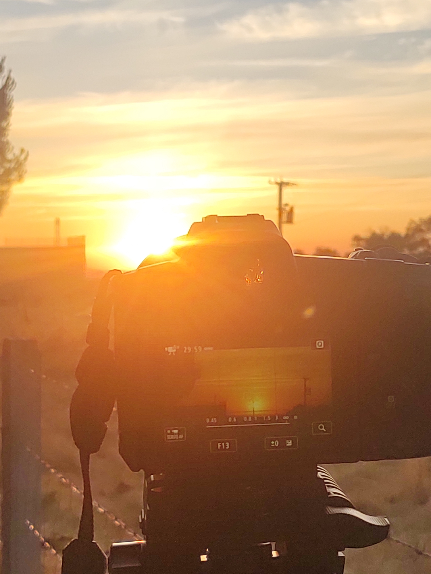 The image shows a close-up of a black digital camera on a tripod, silhouetted against a bright sunset. The camera's LCD screen displays a zoomed-in view of the horizon with the sun partially obscured by clouds, along with camera settings including "29.59", "0.45", "0.6", "0.8", "1.5", "F13", and "+0". In the background, there is a treeline, a utility pole, and some low-lying structures, suggesting an outdoor, possibly rural, setting. The sky is a mix of orange, yellow, and blue from the sunset. The foreground shows a barbed wire fence. There are no people visible in the image. The setting appears to be the municipality of Sobradillo, Spain, given the presence of a rural landscape with a utility pole and structures typical of such an area.