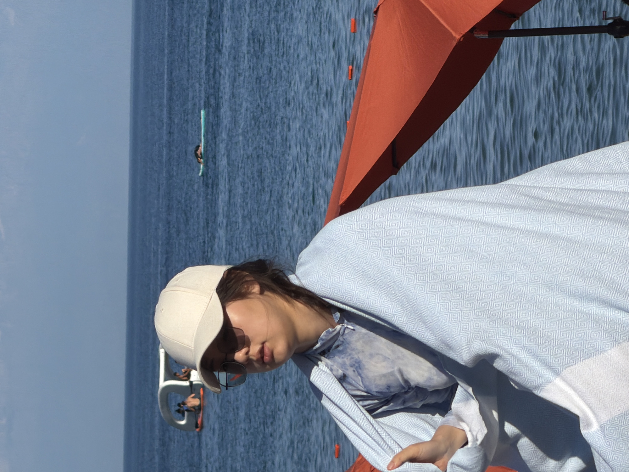 A young woman wearing a tan baseball cap and sunglasses is wrapped in a light blue blanket with a diamond pattern. She is sitting outdoors with a body of water in the background. The water appears to be a lake or ocean, with a clear blue sky above. In the distance, there are a few other people on a white floating structure and one person on a paddleboard. An orange umbrella is partially visible in the foreground on the right side of the image. The woman's clothing visible under the blanket is a tie-dye shirt.