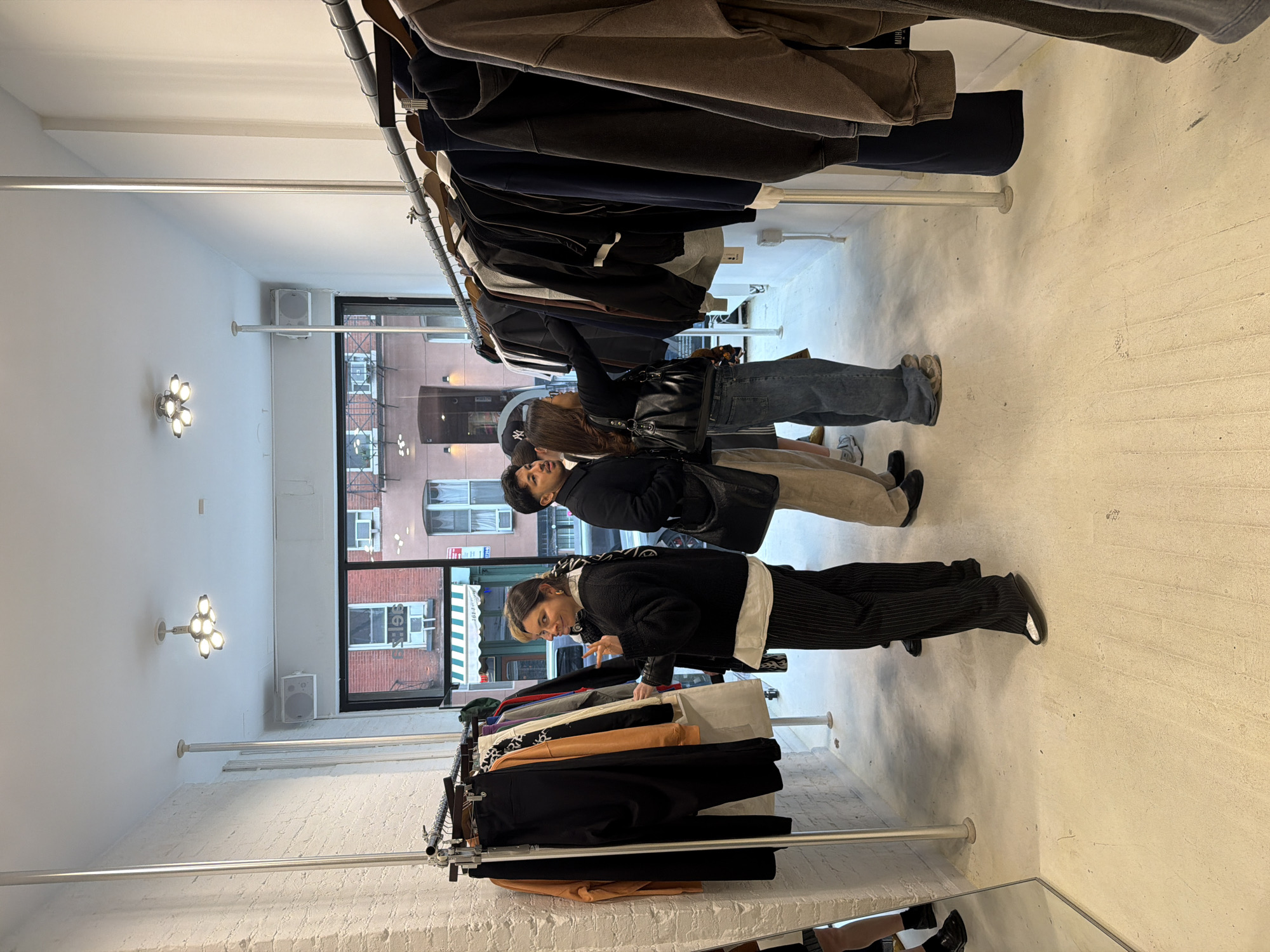 The image shows the interior of a clothing store located in New York City, United States of America. Three people are browsing through racks of clothing. Two clothing racks are visible, one containing dark-colored garments and the other with a variety of colors. The store has white walls, concrete floors, and multiple hanging lights on the ceiling. A window in the back displays a street scene with buildings and a striped awning.