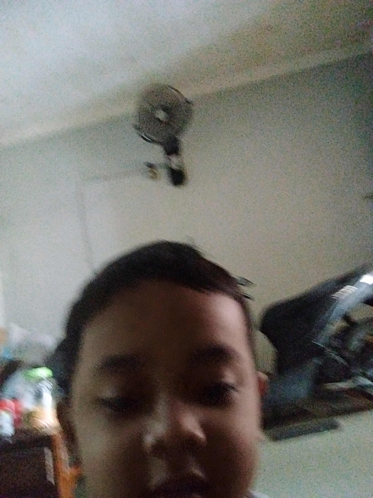 The image is a selfie taken by a young boy indoors, with the camera focused on his face. In the background, a ceiling fan is visible, along with what appears to be a wall and some blurred objects. The lighting is dim, and the image quality is poor due to low resolution and noise. There are no specific landmarks or context to determine the location in Pepelegi, Indonesia.