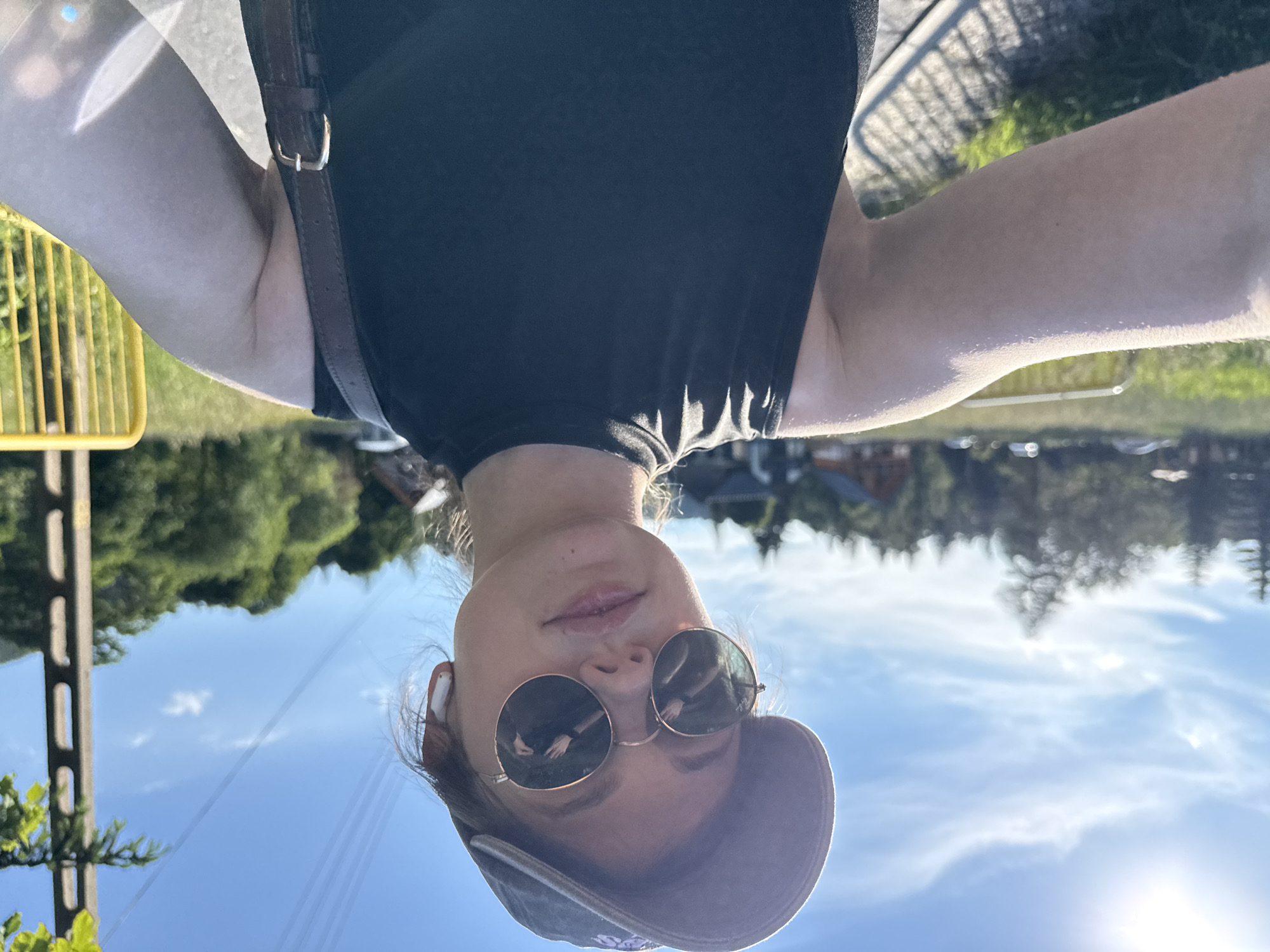 A young woman is captured in a selfie outdoors, wearing a black sleeveless top, a baseball cap, and sunglasses. She has white earbuds in both ears. The background shows a bright blue sky with some clouds and sunlight illuminating the scene. Trees and some buildings are visible in the distance, suggesting a rural or suburban setting. A yellow barrier is present on the right side of the frame. The lighting indicates it is daytime, possibly late afternoon due to the sun's angle. The location is identified as Szklarska Poręba, Poland.