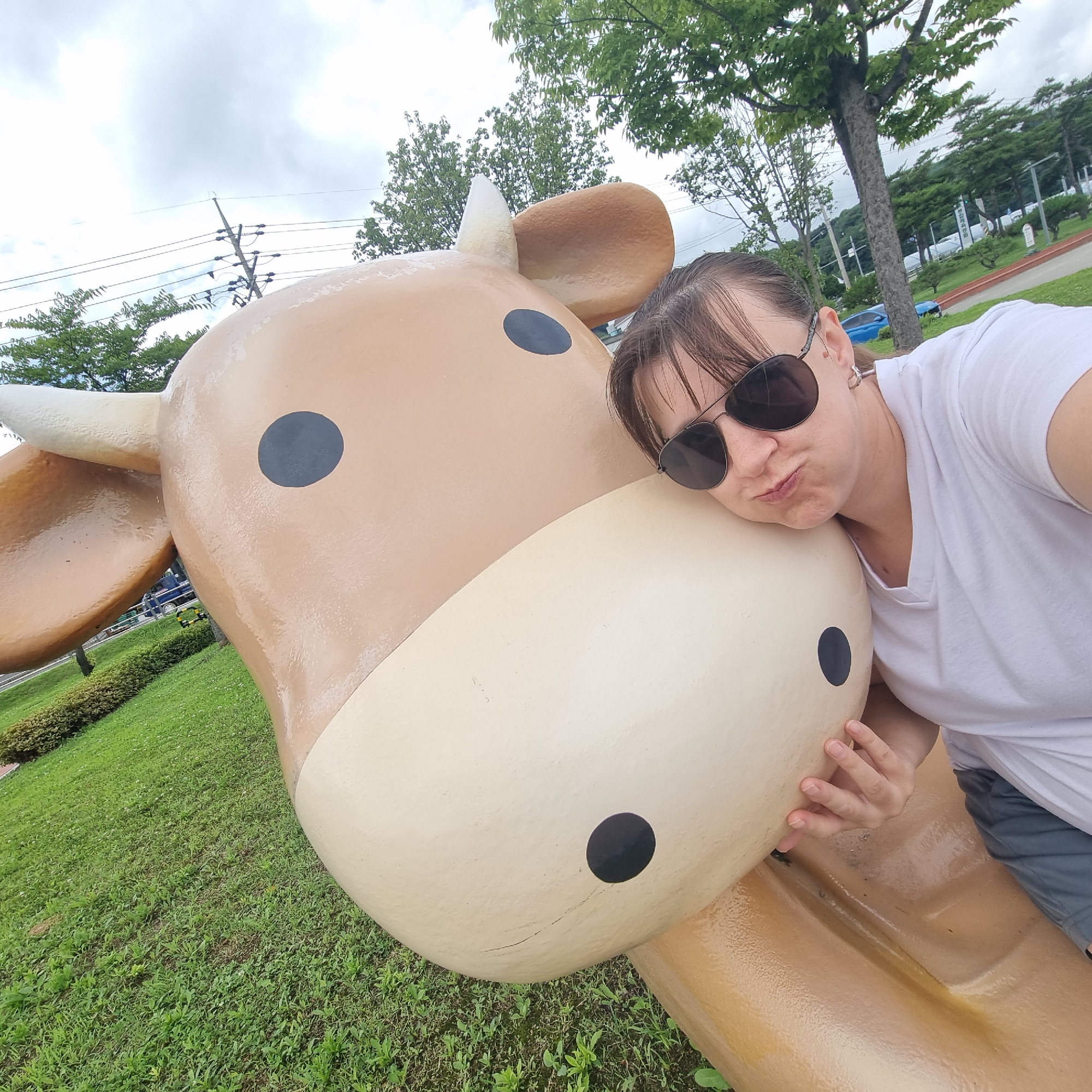 In Dunnae-myeon, South Korea, a woman wearing sunglasses and a white t-shirt rests her head on the cheek of a large, cartoonish cow sculpture. The sculpture is light brown and beige with black circular eyes. The woman has her eyes closed and appears to be posing. The scene is outdoors, with a grassy area and trees in the background, under a cloudy sky. Power lines are visible against the sky.