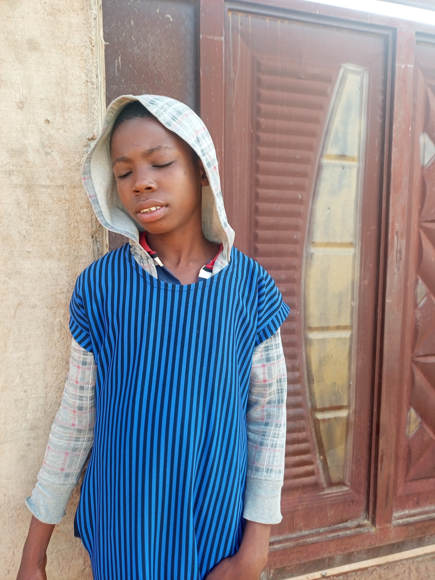 A young boy stands with his eyes closed, leaning against a textured, light-colored wall. He wears a hooded garment with a light gray hood lined with a red and blue checkered pattern. His outer shirt is a blue and black vertical striped tunic, with light gray sleeves that have a subtle checkered pattern in red and blue. The boy's mouth is slightly open, revealing his teeth. To his right, a large, dark reddish-brown door with horizontal slats and a decorative, elongated pane of light-colored material is visible. The setting appears to be outdoors, possibly in a residential area of Tempure, Nigeria, indicated by the architecture and the natural lighting.