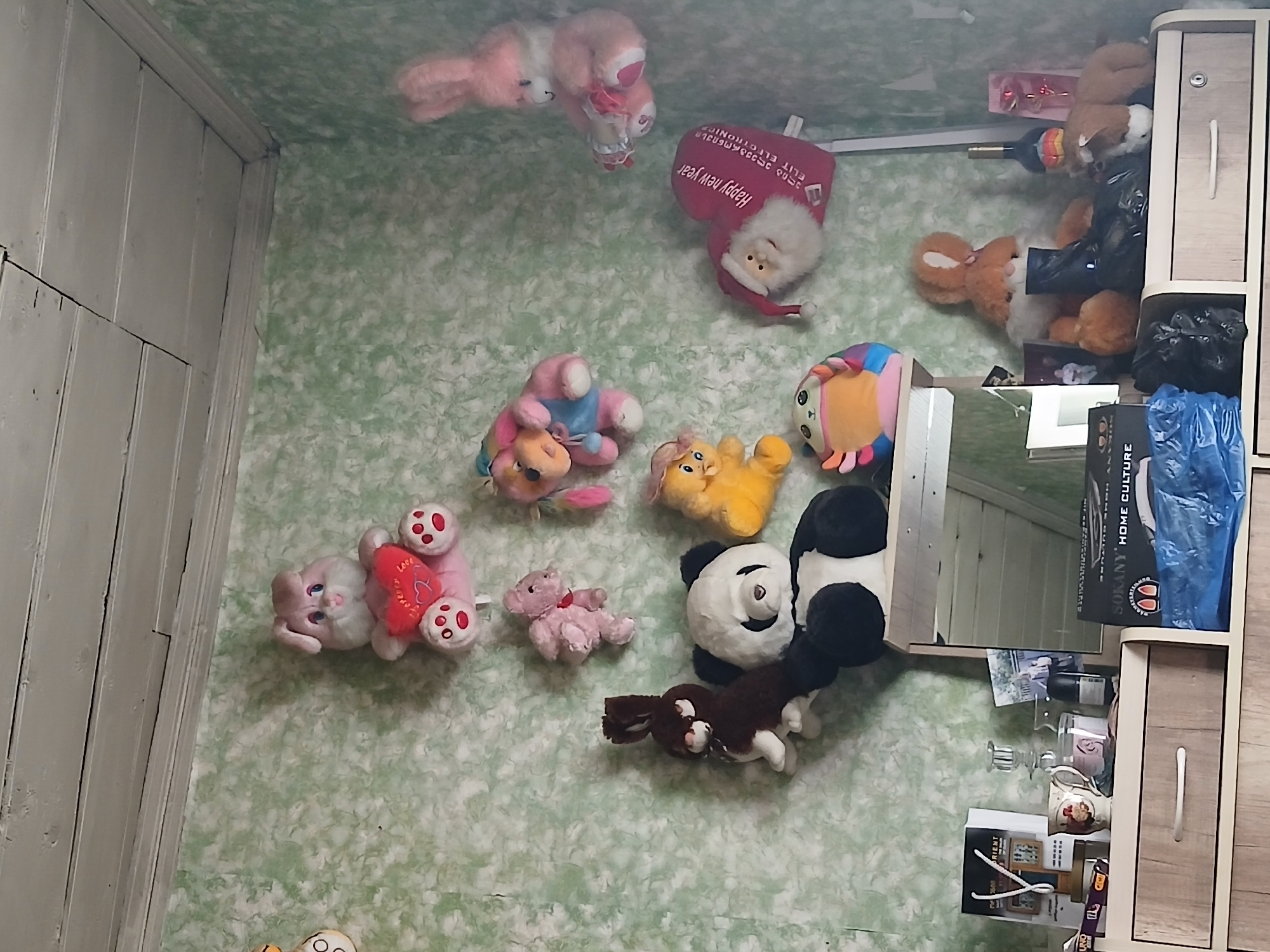 In a dimly lit room with light green, textured wallpaper and a wooden ceiling, a collection of stuffed animals is displayed on a wall and atop a wooden dresser. The stuffed animals include a large pink bunny holding a red heart, a small pink teddy bear, a pink and blue dog-like creature, a yellow plush toy, a brown and white bunny, a panda, a colorful crab-like creature, and a large pink bunny. A heart-shaped pillow with an image of Santa Claus and text in Georgian reads "Happy New Year". On the dresser, various items are visible, including mugs, candles, a bottle of wine, a decorative rose, and a small stuffed horse. Also on the dresser, there are blue and black plastic bags and several boxes, one of which has the text "SOKANY HOME CULTURE". The scene appears to be an indoor residential setting. There is no indication of the city of Telavi, Georgia, or any specific location within it.