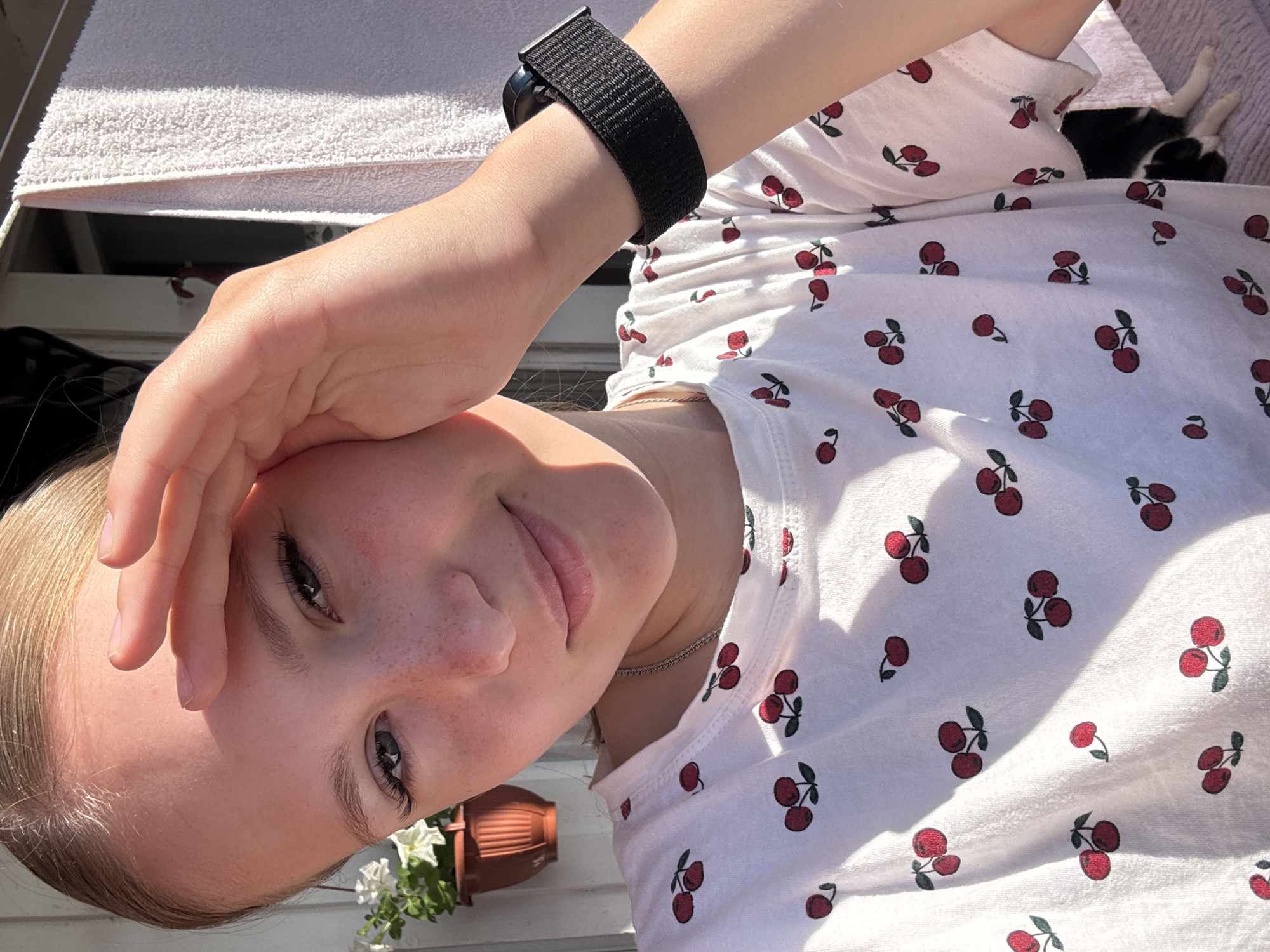 A young girl with blonde hair pulled back is positioned in the foreground, her face turned towards the camera. She is wearing a white t-shirt with a repeating pattern of red cherries. Her right hand is raised, resting on her forehead, obscuring part of her hairline. Sunlight casts a shadow across her forehead and cheek. In the background, a white towel is hanging, and to the left, a potted plant with white flowers is visible. The scene appears to be outdoors, possibly on a balcony or porch, with a wooden structure behind the girl.