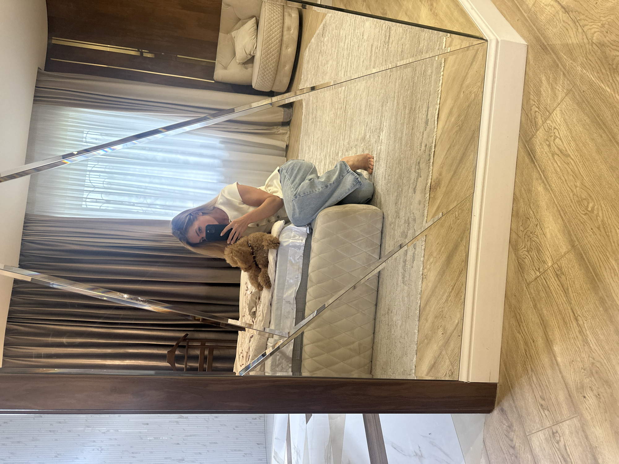 The image shows a mirror reflection of a room interior. A woman is lying on a bed taking a picture with her phone. A small brown poodle sits next to her. The bed has a beige, quilted headboard. Behind the bed are brown curtains and a dark brown wooden headboard with gold accents. Reflected in the mirror is a beige rug and a cream-colored sofa. The room has light wood flooring and white trim around the mirror. The bottom of the mirror reflects a white brick wall and marble flooring. There is no evidence to indicate this photograph was taken in the city of Armavir, Russia.