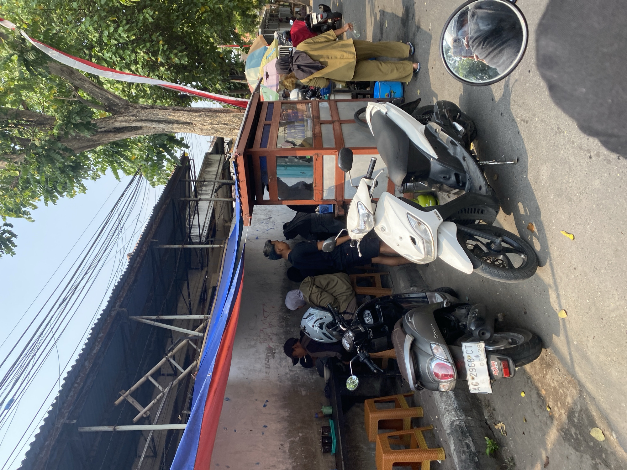 In Kediri City, Indonesia, a street scene unfolds in front of an outdoor eatery. Two motorcycles, a dark grey Honda Scoopy with license plate AC 2966 CT and a white Yamaha, are parked on the asphalt road. Several people are seated at a long table under a sheltered awning, some eating, others talking. The eatery has a small, open-air stall with glass windows displaying goods and a cashier. To the right, a woman in a tan hijab and matching pants stands near the stall, holding a clear plastic bottle. A tall tree with a red and white Indonesian flag draped from its branches stands to the right of the eatery, amidst other greenery and a blue sky. The overall setting suggests a typical day at a local food vendor.