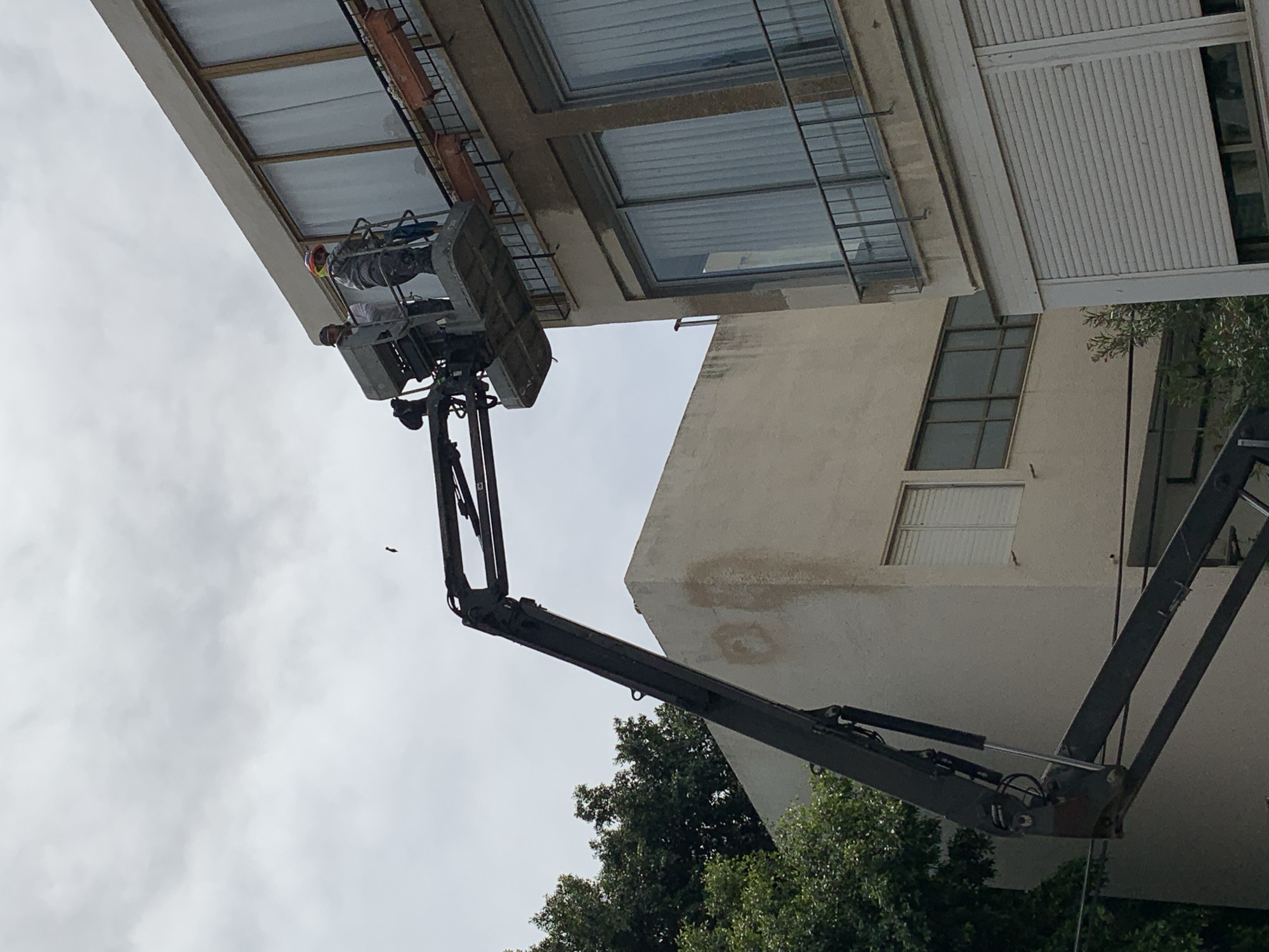 A hydraulic lift is elevated to a multi-story building in Tel-Aviv, Israel. A construction worker in a hard hat operates from the basket of the lift at the building's exterior. The building is a light, off-white color with visible windows. Trees partially obstruct the lower portion of the lift. The sky is overcast.