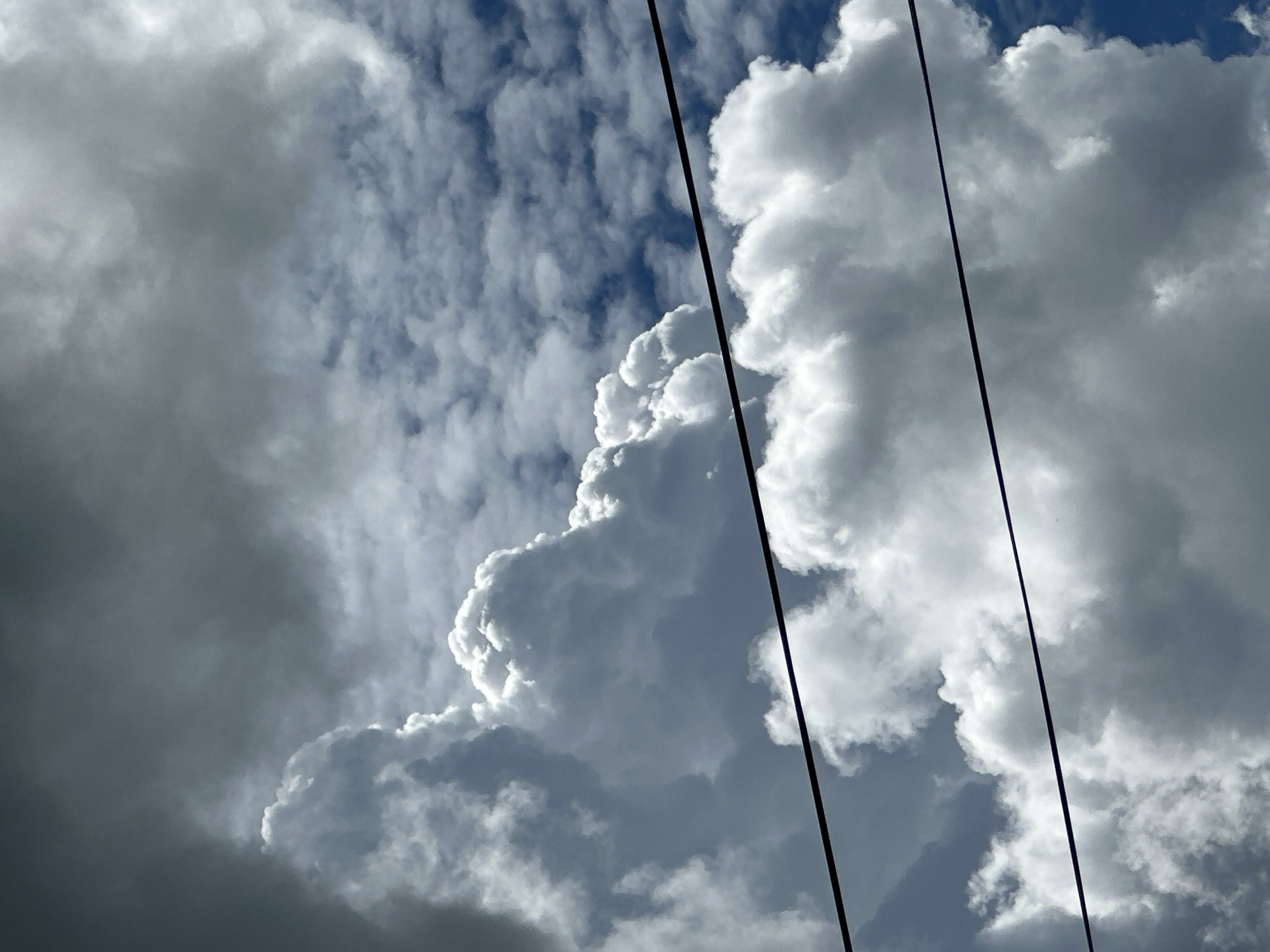 This is a close-up, upward-facing shot of a dramatic sky filled with a variety of clouds. The main subjects are the clouds themselves, ranging from dark, heavy storm clouds on the left to bright, billowing cumulus clouds illuminated by sunlight on the right. The sky is visible in patches between the clouds, showing a deep blue.
Two thin, black lines, likely power lines, run diagonally across the frame from the top left to the bottom right, partially obscuring the sky and clouds.
The light suggests it is daytime, with the contrast between the shadowed and sunlit clouds indicating a dynamic weather system. The clouds' formations hint at potential rain or a storm approaching or passing. The scene evokes a sense of natural power and the vastness of the sky. There is no visible text in the image.