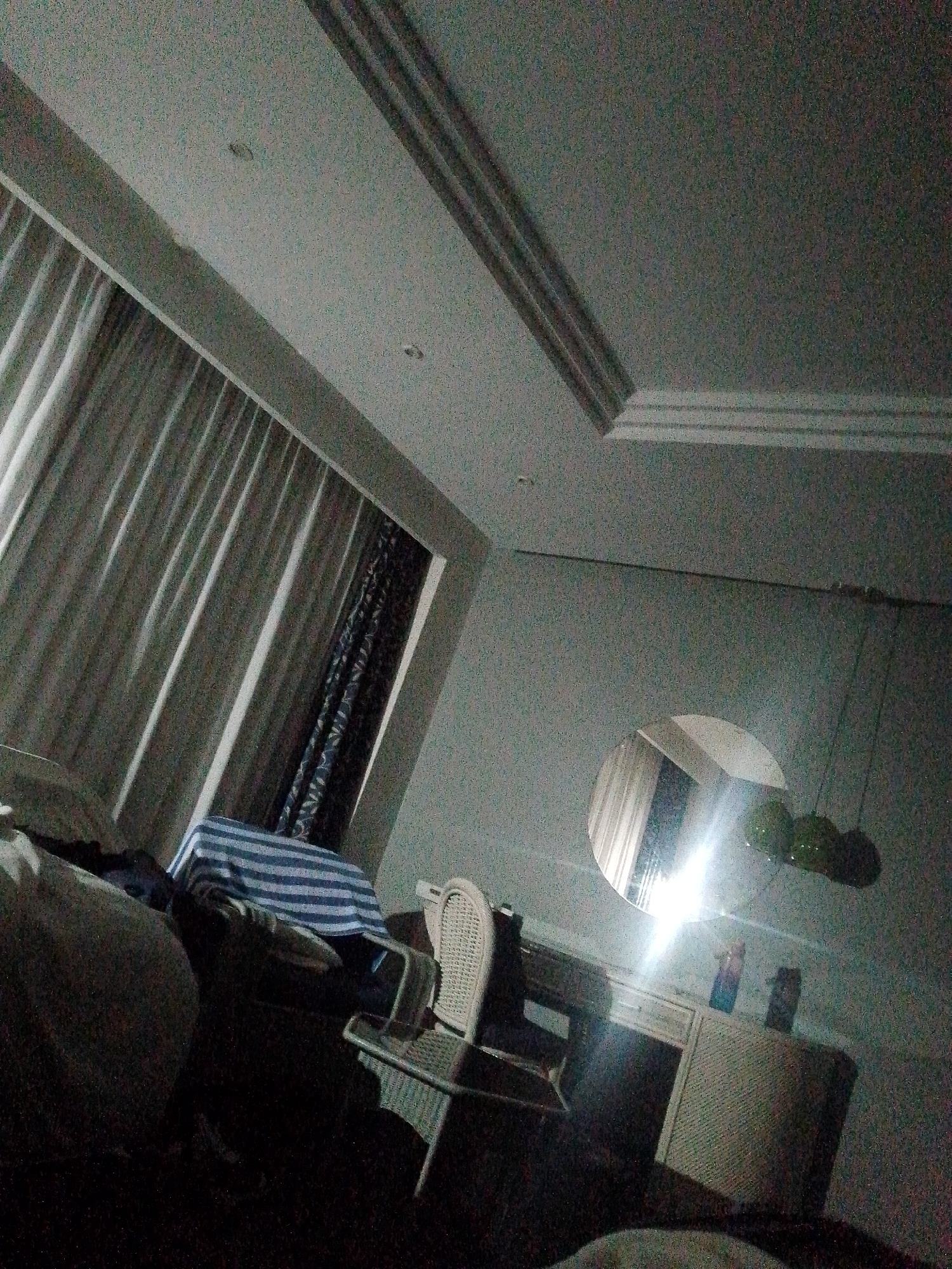 The image depicts a dimly lit interior, possibly a bedroom. A large window with layered curtains is on the left side of the frame. A bed is partially visible in the foreground. A desk with a mirror and chair is in the middle of the room. There are a few decorative items on the desk. Three hanging decorative spheres are on the right side of the frame. The location is given as Pedro González, Venezuela. No people are visible.