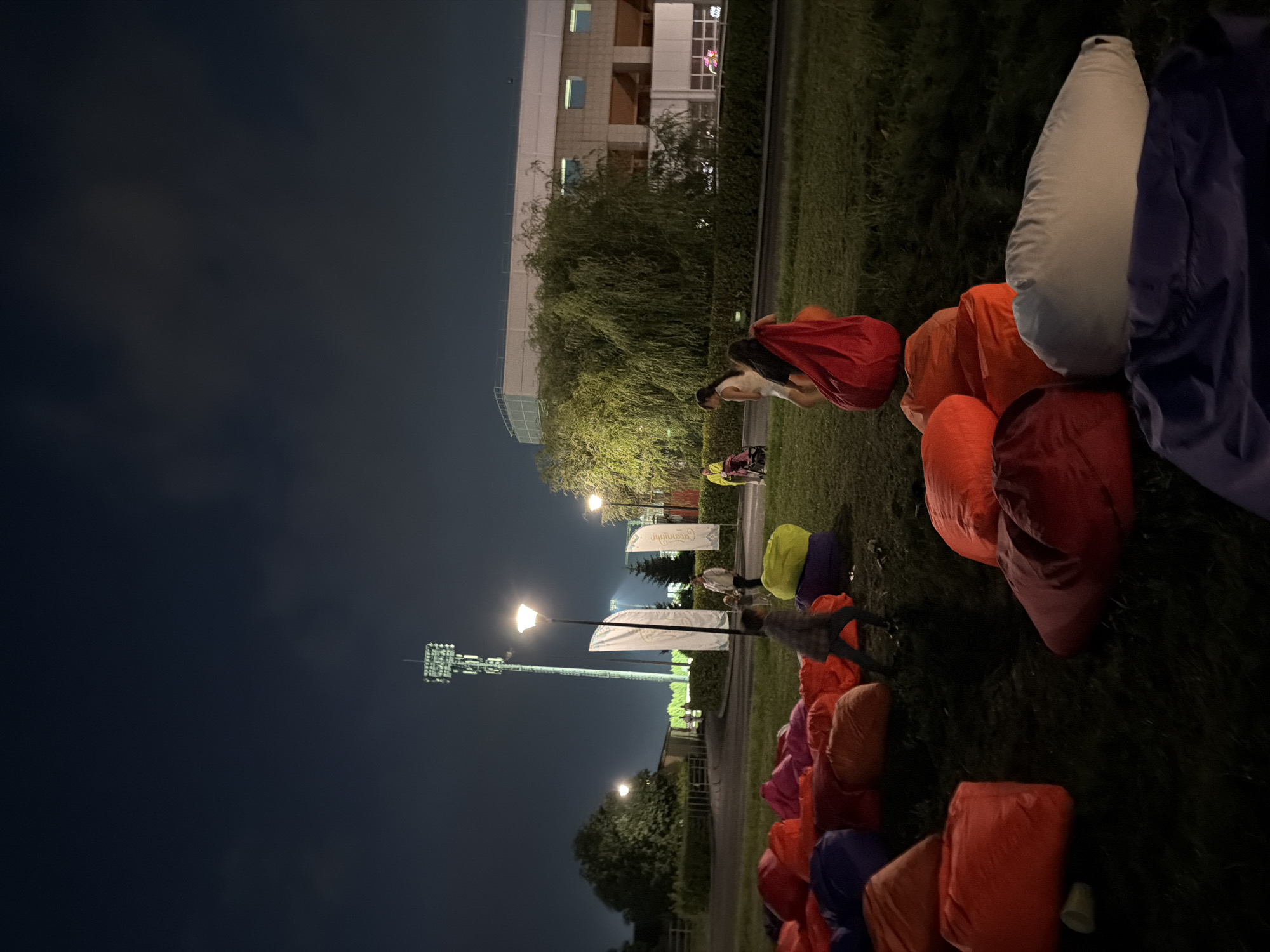 At night, a group of people are gathered in a park. Beanbag chairs of various colors are scattered on the grass. In the background, a tall tower with lights is visible, along with a building. Several people are walking around the beanbags, some are carrying them. A woman with a stroller is also present. The overall scene suggests an outdoor event or gathering in a park.