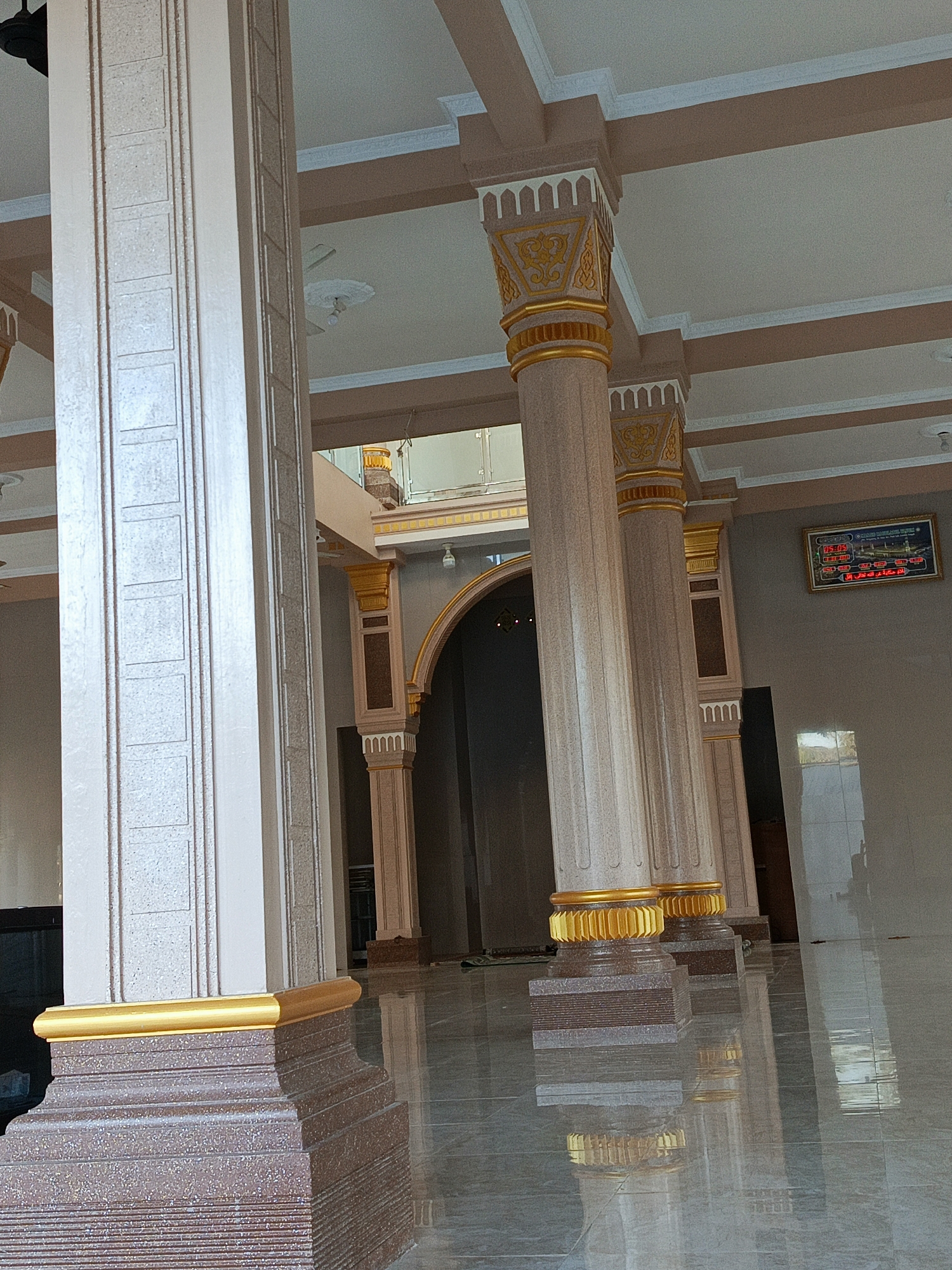 The image shows the interior of a mosque in Kuta, Indonesia. The scene is characterized by large, ornate pillars with gold accents. The floor is highly reflective, showing a mirror image of the pillars and the surrounding architecture. In the background, an arched doorway is visible, leading to a darker area. The ceiling features recessed lighting and decorative molding. A digital display board showing prayer times is mounted on the wall to the right, with Arabic text and numerals displayed in red. The lighting suggests it is daytime, with natural light filtering in, though the exact time of day is not discernible. There are no people visible in the frame. The overall impression is one of grandeur and religious solemnity.
