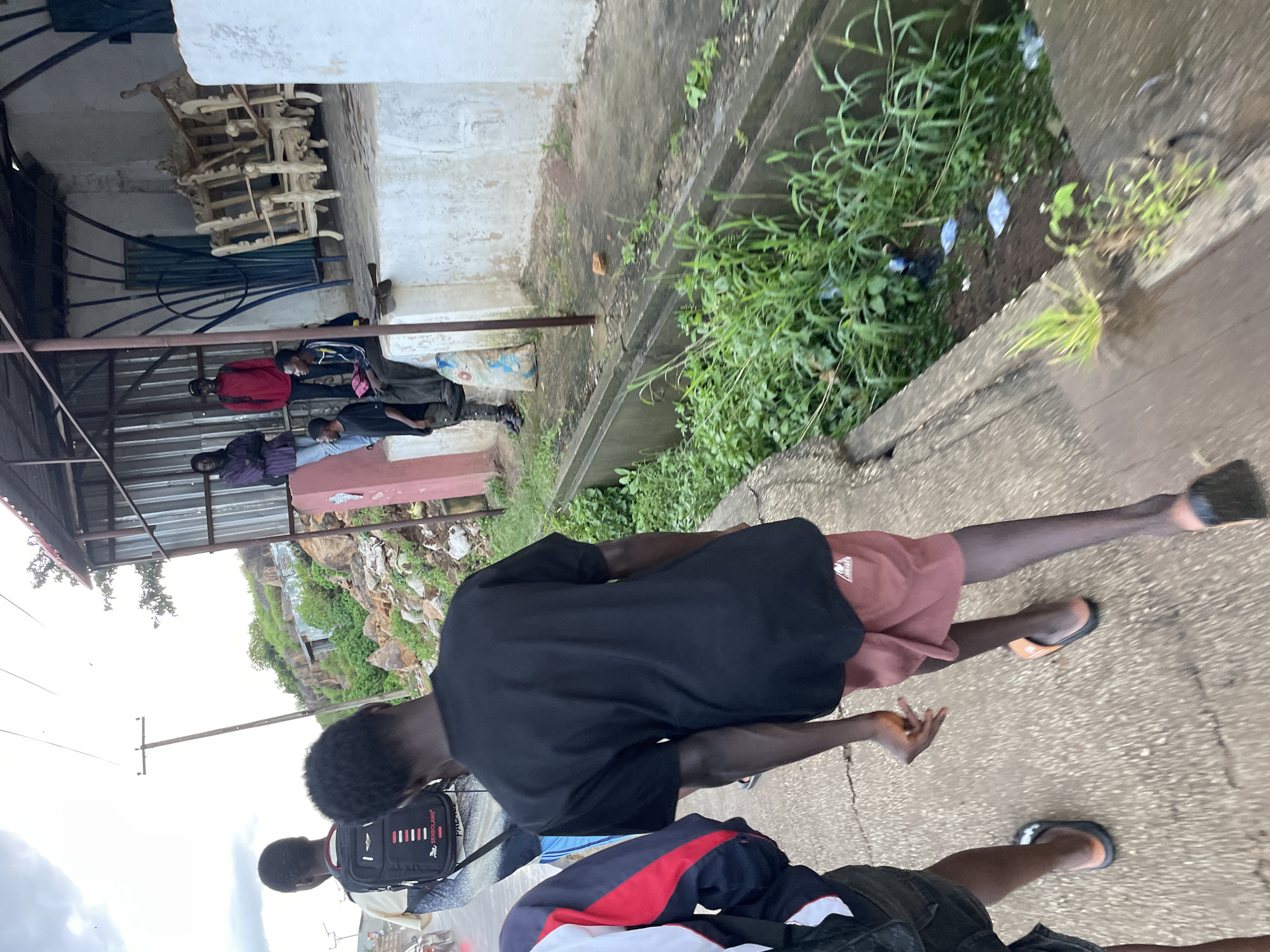 The image captures a street scene in Jos, Nigeria, on an overcast day. In the foreground, two young Black males are walking away from the camera, with the one on the right wearing a black t-shirt and reddish-brown shorts, and the one on the left partially visible in a dark jacket with red and white accents. The male on the right has a distinctive hand gesture. They are walking on a cracked concrete surface next to a drainage ditch filled with lush green vegetation and some scattered litter.
Further back, under a corrugated metal roof supported by metal poles, several individuals are gathered. One person in a purple shirt is standing with arms crossed, while another in a red shirt stands nearby. A man in a blue and black jacket is seated on a ledge, and another individual in a dark shirt is also present. Behind them, ornate wooden furniture is stacked, possibly awaiting sale or repair. The background reveals a rocky hillside with some structures and sparse vegetation, suggesting an urban or semi-urban fringe environment. The overall impression is one of a typical street scene in a Nigerian city, with elements of commerce, community gathering, and everyday life.