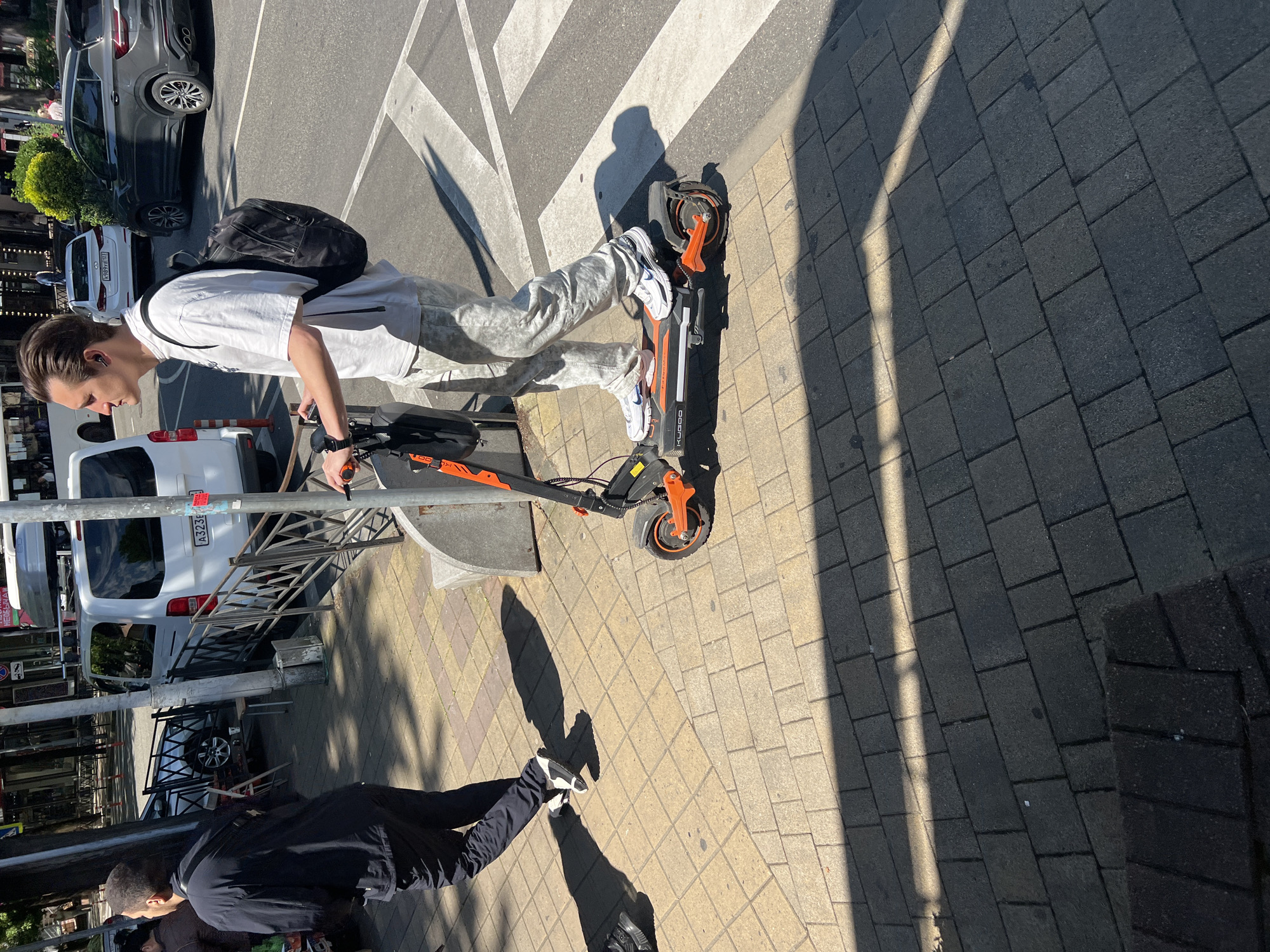 The image captures a sunlit urban street scene in Sochi, Russia, featuring two men and an electric scooter. The main subject, a young man with dark hair slicked back, is prominently featured on the right side of the frame. He wears a white t-shirt, light gray patterned pants, and white sneakers, with a black backpack slung over his shoulders. He has earbuds in his ears and a watch on his left wrist. He is standing beside an orange and black electric scooter, with his right hand resting on its handlebar. The brand "KUGOO" is visible on the scooter's deck.
The man stands on a light-colored paved sidewalk, which runs alongside an asphalt road featuring a white zebra crossing. In the background, several vehicles are visible, including a dark grey SUV and a white cargo van. The white van's license plate clearly displays "A323КХ 93".
Another man, dressed in a dark jacket and pants with a dark backpack, is partially visible in the lower left of the frame, walking away from the camera on the same sidewalk. Both men and the scooter cast long, sharp shadows on the ground, indicating bright sunlight and clear weather, likely mid-morning or late afternoon. Metal barriers and a street pole are also part of the street furniture in the scene. The overall impression is a casual, active moment in a city environment.
