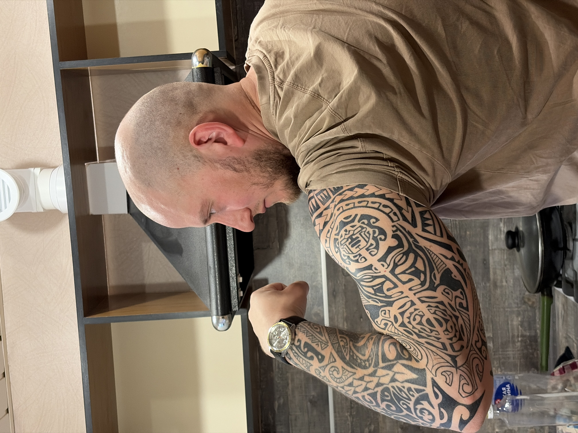 An image, rotated 90 degrees clockwise, captures a man with a bald head and a short beard, seen from the mid-chest up. He wears a casual, short-sleeved, tan-colored t-shirt. His head is tilted forward and down, eyes cast downwards, making his expression unreadable. His left arm is bent at the elbow, with a loosely clenched fist, and is heavily tattooed with intricate black tribal/Polynesian designs covering the forearm and upper arm. A wristwatch with a metallic casing and a dark strap is worn on his left wrist.
The setting appears to be indoors, likely a room with shelving units. Dark wooden or metal shelves with lighter beige/brown interior panels are visible in the background, extending vertically. To the left, a white PVC ventilation pipe and a circular vent cover are attached to a light-colored wall or cabinet. A dark, possibly wooden, floor or countertop with a visible grain texture is beneath his arm. A black metal pole with a shiny chrome-colored top is seen behind his head, suggesting a support structure for shelving or equipment. In the bottom right corner, partially obscured, is a pot or pan lid with a black handle and a clear plastic bottle with a blue label. Visible Cyrillic text on the bottle label includes "ВЯТ" and other characters, possibly "Вятская". The overall lighting suggests an indoor, artificially lit environment.