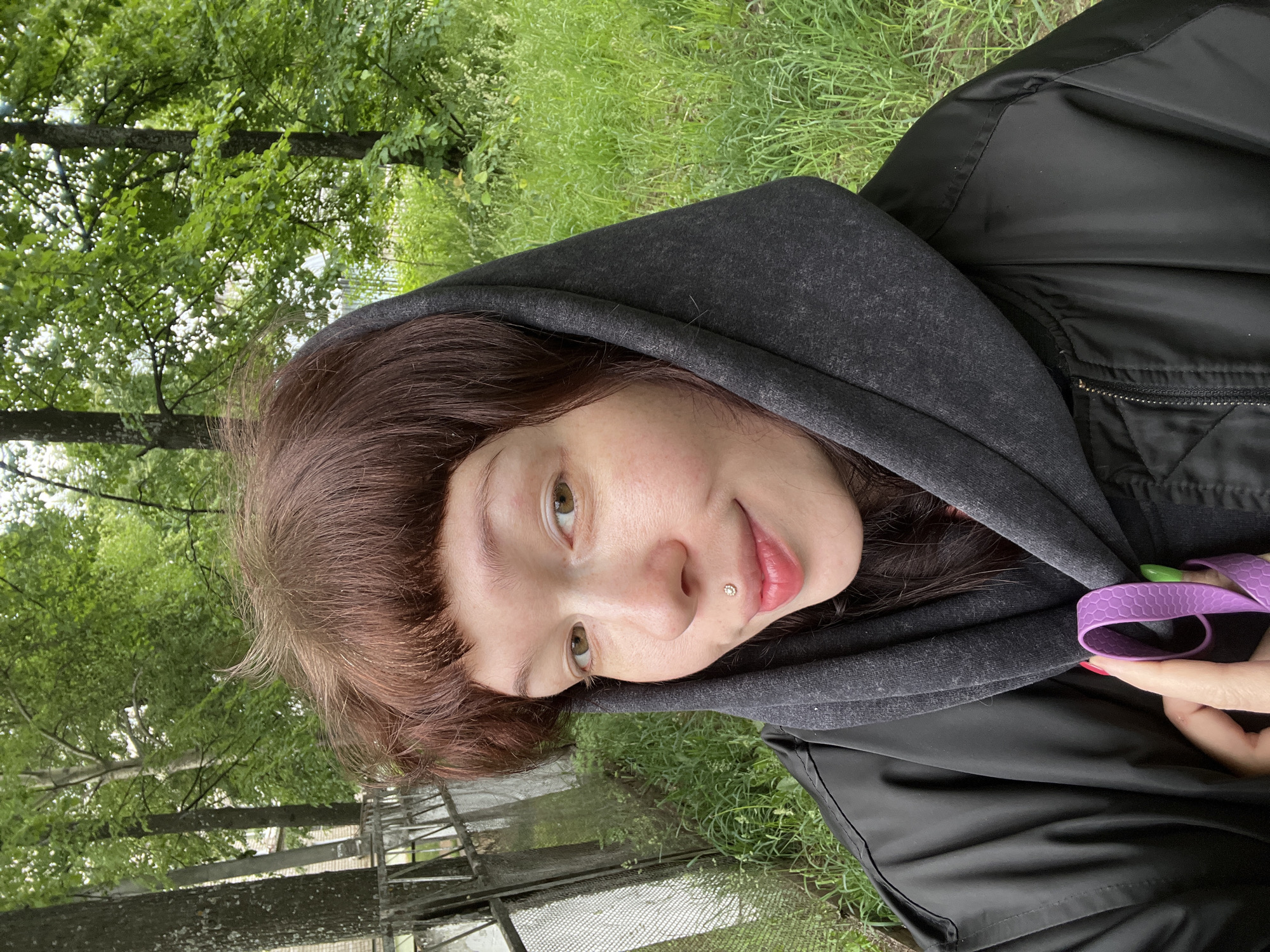 A close-up, selfie-style portrait features a young woman in an outdoor setting in Chaikovsky, Russia. She has shoulder-length reddish-brown hair with bangs that fall across her forehead. Her light-colored eyes look directly at the viewer with a calm, pleasant expression, accompanied by a subtle smile. A small, shiny labret piercing is visible just below her lower lip.
The woman is dressed in a dark grey hooded sweatshirt layered under a black jacket. Her hand, visible in the lower right, holds a textured, bright purple strap or band. Notably, her fingernails display a playful style, with one painted bright green and another red.
The background is a vibrant display of nature, dominated by lush green trees with dense foliage and bright green grass, suggesting a park or natural area. A metal fence or railing is partially visible in the mid-ground. The natural, diffused light indicates it is daytime, likely on an overcast day or in a shaded spot under the trees. There is no visible text.