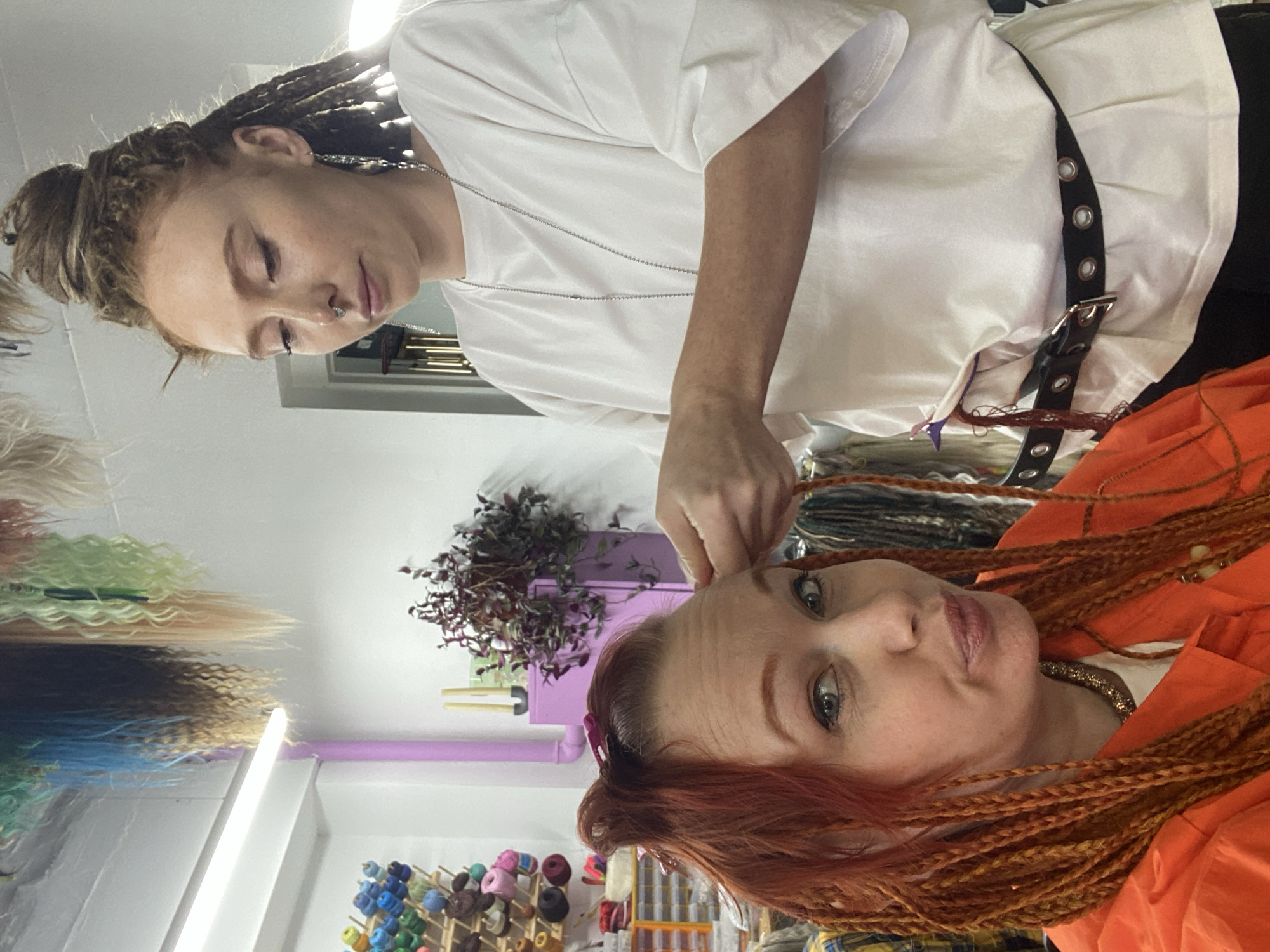 The image, rotated 90 degrees clockwise from its natural orientation, depicts an indoor scene within a hair styling studio or salon in Sochi, Russia.
Two women are the central figures. One woman, with light brown dreadlocks pulled back into a bun and a nose piercing, wears a white short-sleeved t-shirt and a black belt with silver eyelets. She is positioned to the left (when viewed as correctly oriented) and is focused intently on the hair of the second woman, indicating she is the stylist.
The second woman, with vibrant reddish-orange hair styled in numerous long braids or dreadlocks, looks directly at the camera with a slight pout or smirk. She is wearing an orange collared shirt and a visible gold-toned necklace. Her intricate braids are prominent, falling over her shoulders and chest.
The primary activity is hair styling, with the first woman meticulously working on the braids or extensions of the second woman. The background features shelves holding a variety of colorful spools, possibly yarn or hair extension materials, along with other craft or beauty supplies. A small leafy plant in a purple pot is also visible. The space is brightly lit by an overhead strip light, suggesting a functional, professional environment. No discernible text is visible in the image. The expressions convey concentration from the stylist and a self-aware, perhaps playful, demeanor from the client.
