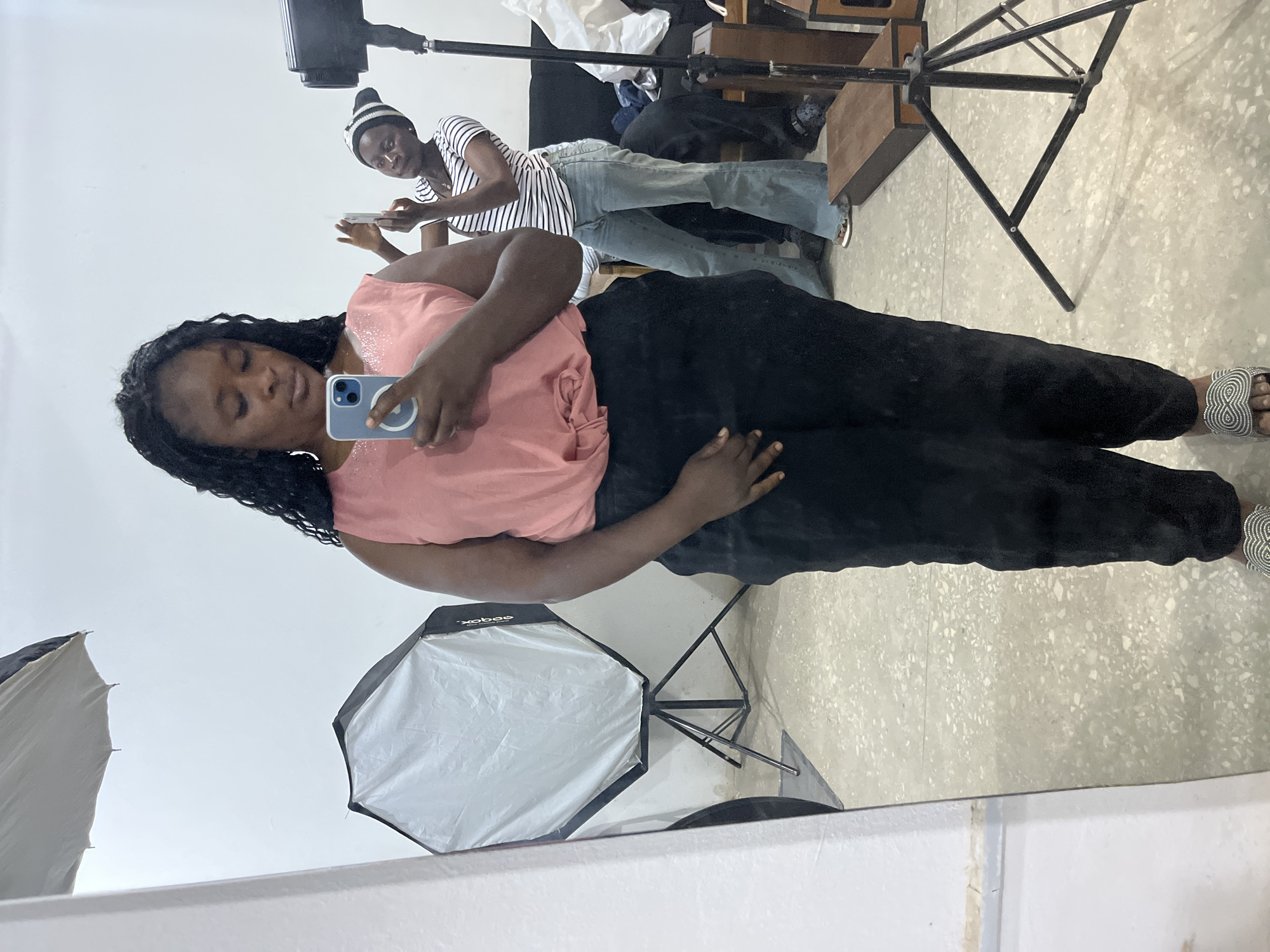 The image shows a woman standing in front of a mirror, taking a selfie. She is wearing a pink sleeveless top and black pants. Her hair is long and braided. In the background, another woman is visible, also taking a photo with her phone. There are several photography lights and equipment in the room, suggesting it might be a studio. The floor is concrete with a speckled pattern. The overall setting appears to be an indoor space with white walls. The location is Gura Top, Nigeria.